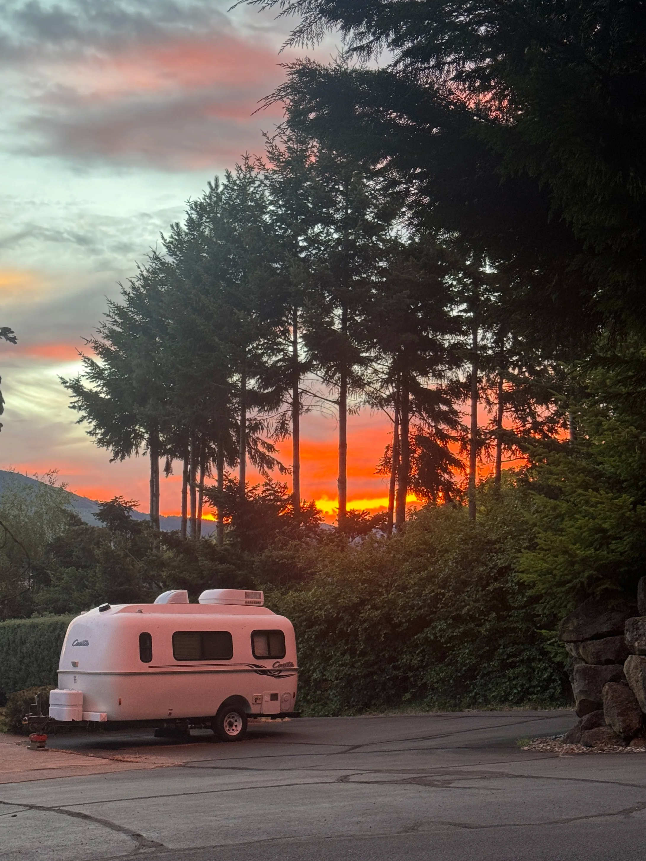 A pink camper trailer parked in a lot with trees and a sunset in the background. The sky has clouds with shades of pink, orange, and purple.