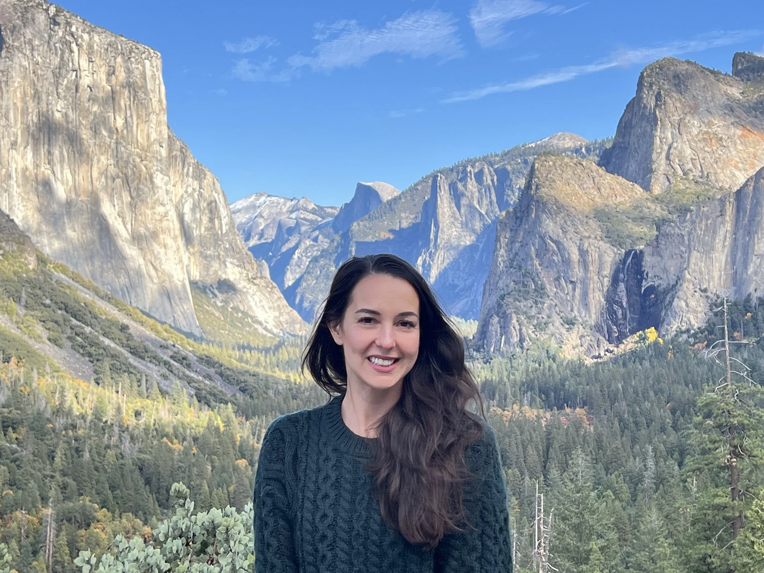 A woman with dark brown hair smiling outdoors in front of a mountain range with rock formations and a blue sky in Yosemite National Park with El Capitan in the background.