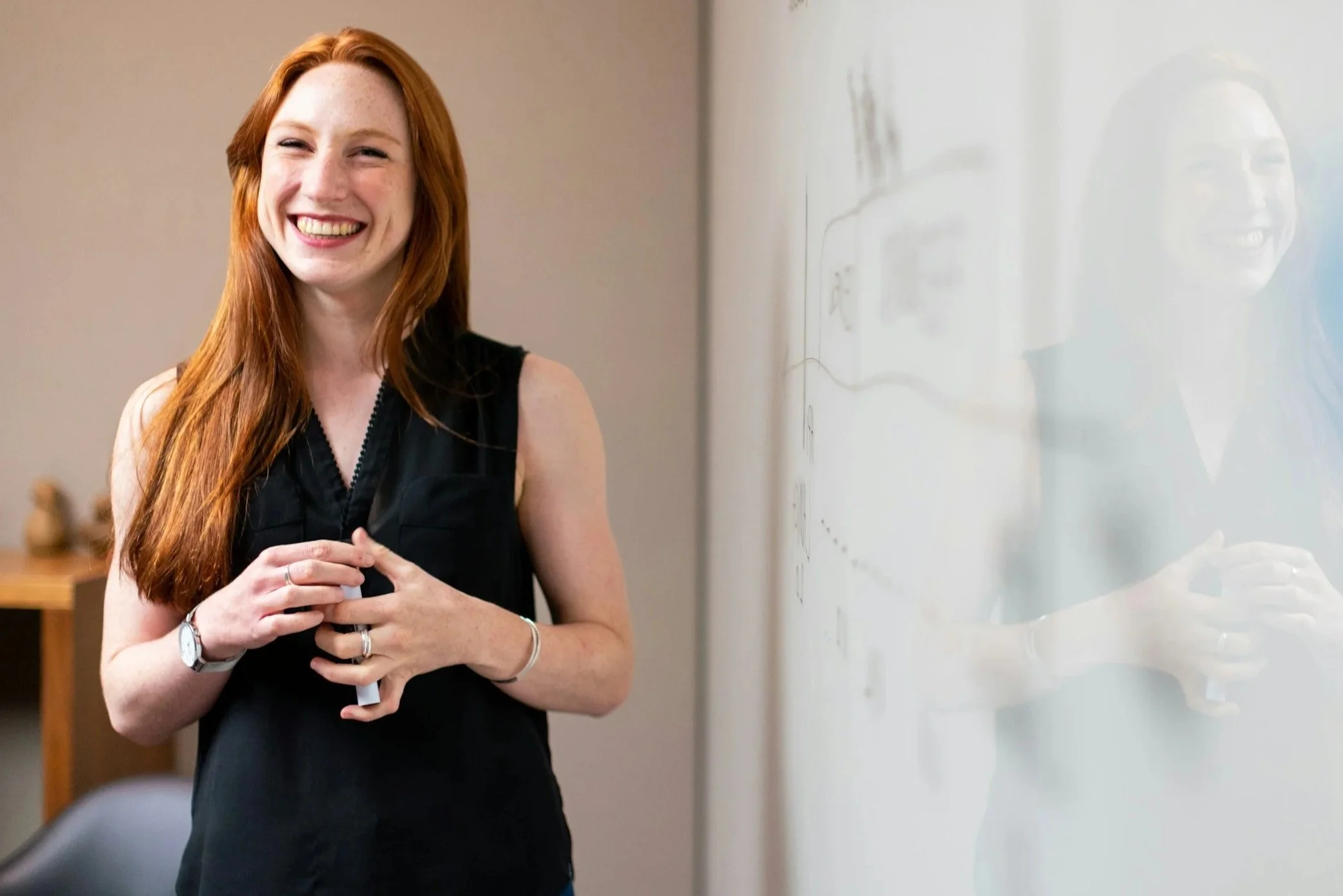 A woman with long red hair smiling and standing in front of a whiteboard, holding a marker in her hands, wearing a black sleeveless top.