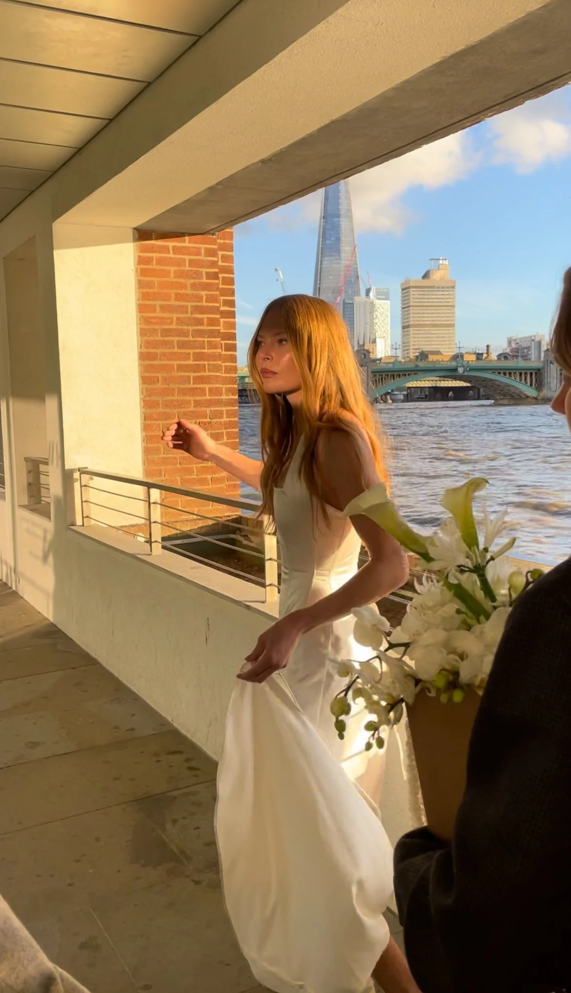 A woman in a white dress standing near a balcony overlooking a river with a city skyline, including a tall modern skyscraper, in the background. Another person is partially visible holding a bouquet of white flowers.