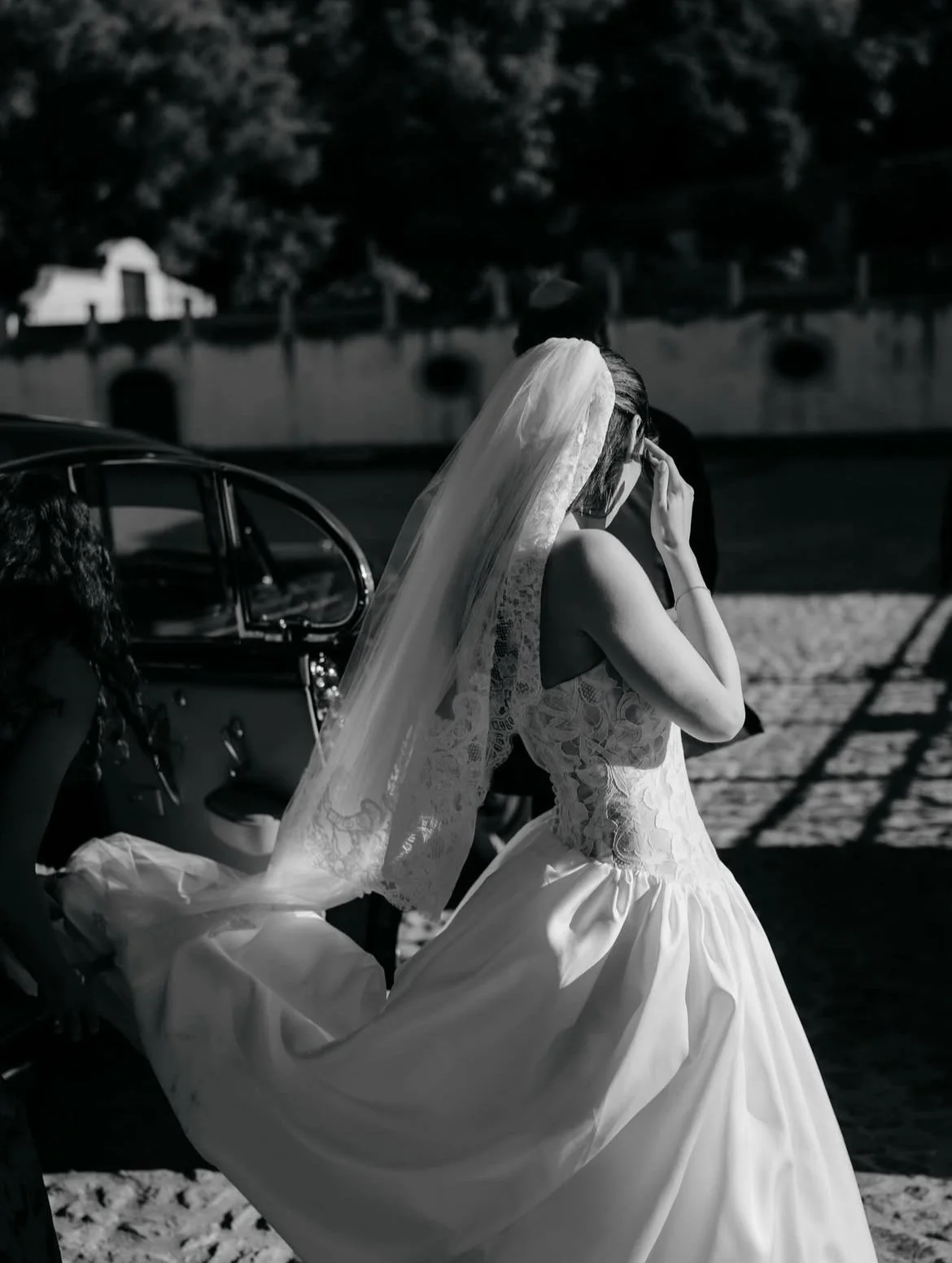 Black and white photo of a bride in a wedding dress with a lace bodice and full skirt, wearing a veil, standing near a vintage car. The bride is touching her face, possibly wiping away tears.