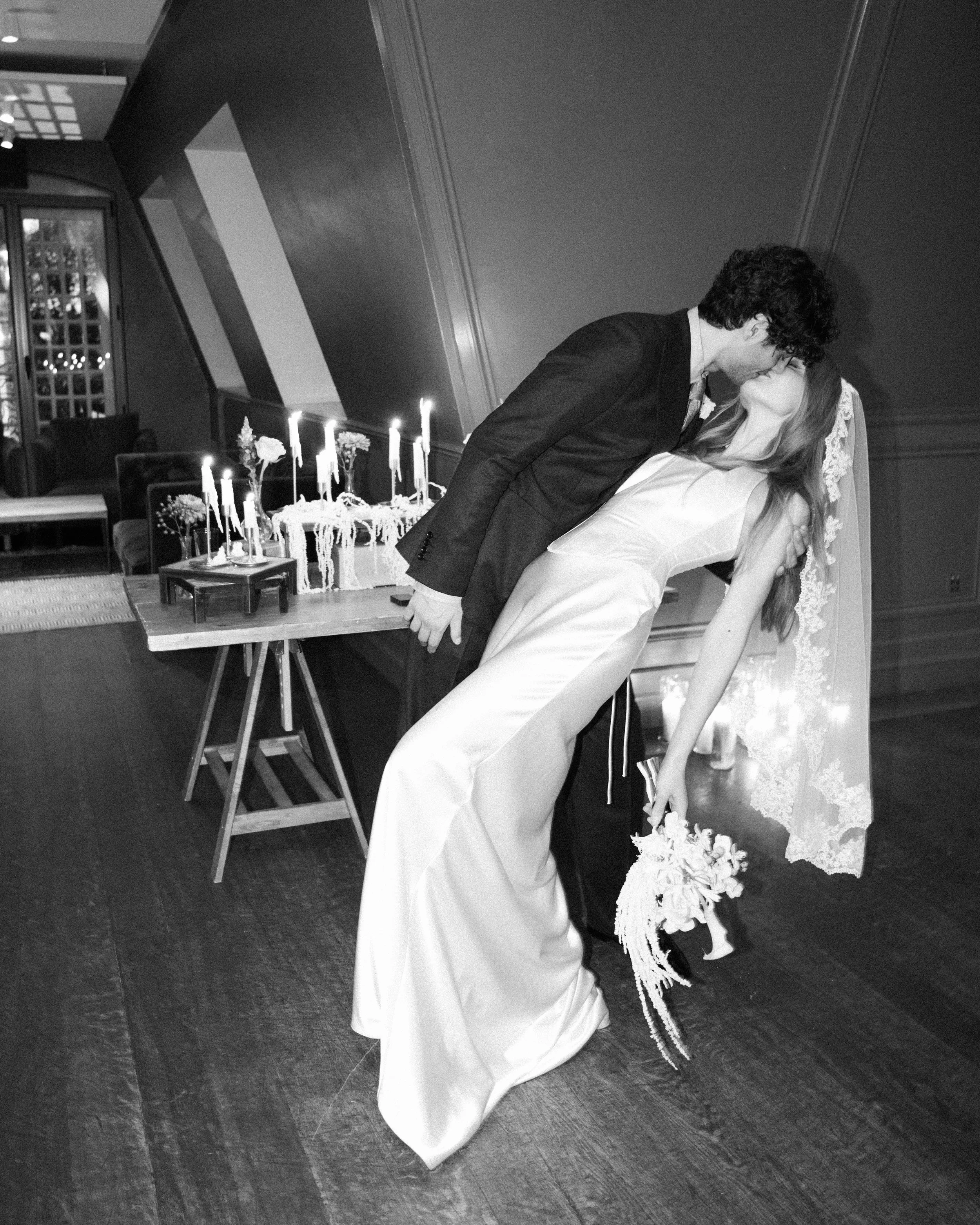 A couple sharing a kiss during a wedding reception. The groom is leaning over the bride who is in a wedding dress, and they are in a decorated room with candles, flowers, and a cake in the background.