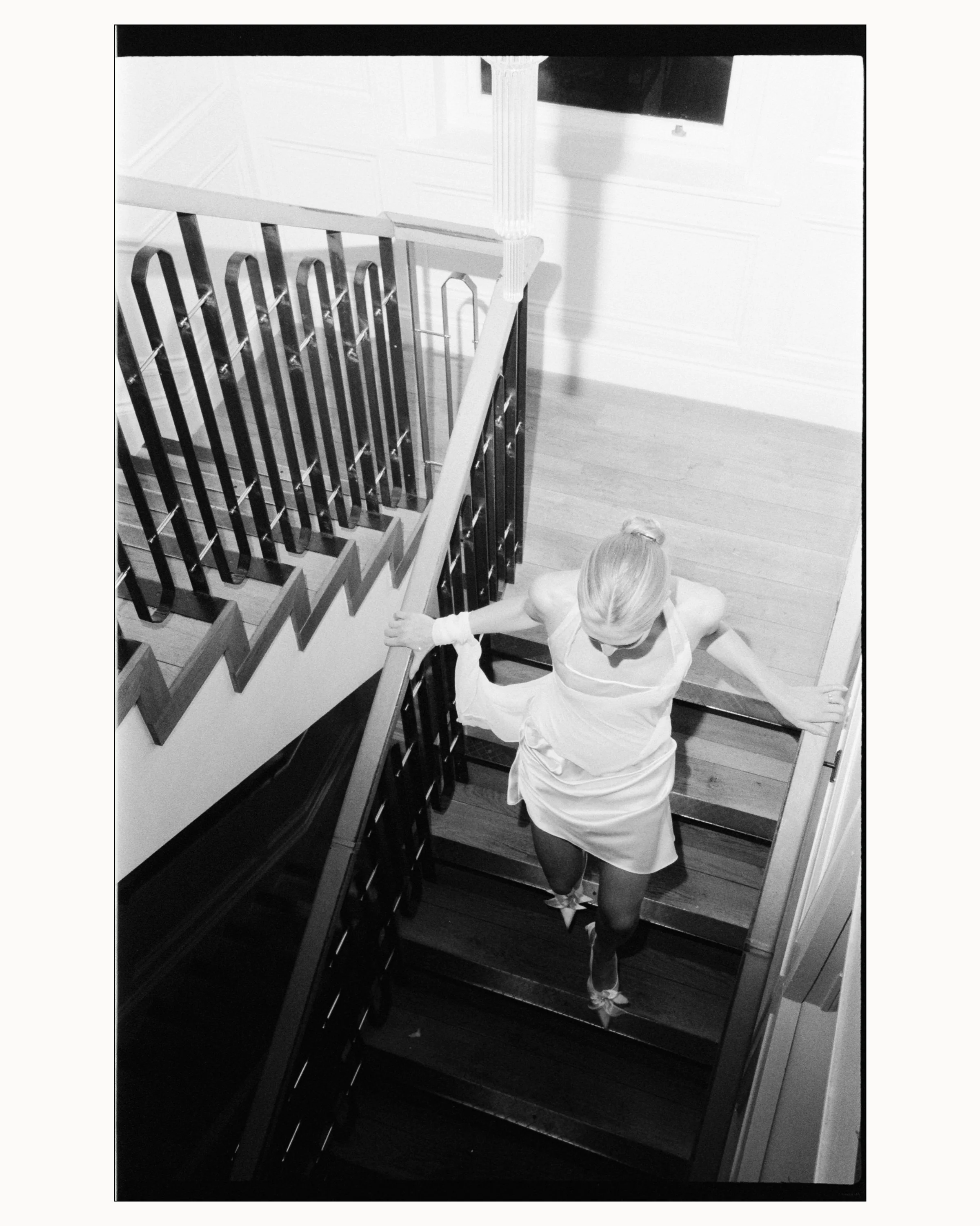 A woman with blonde hair in a bun, dressed in a white dress, is walking up a staircase with dark wooden steps and a white railing, while holding onto the railing with her right hand.