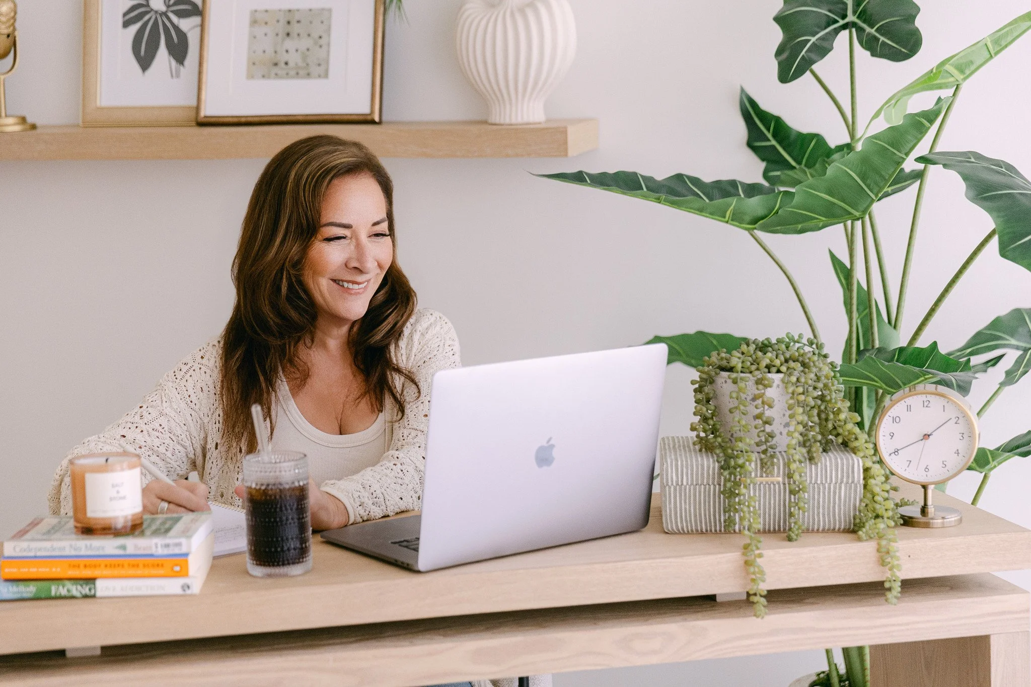 A woman with long brown hair, smiling, sitting at a wooden desk with a laptop, in a room with light-colored walls, decorated with framed artwork, a large potted plant, a white vase, a clock, and books, with coffee cups on the desk.