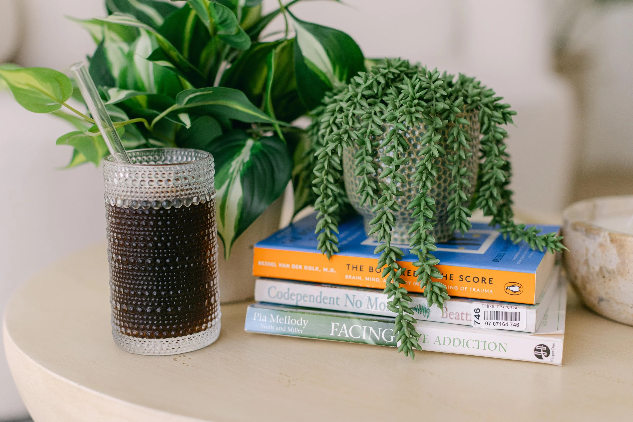 A table with a tall glass vase with a textured dot pattern holding a green leafy plant, and a stack of colorful books, including 'The Body Keeps the Score,' 'Codependent No More,' and 'Facing Love Addiction.'