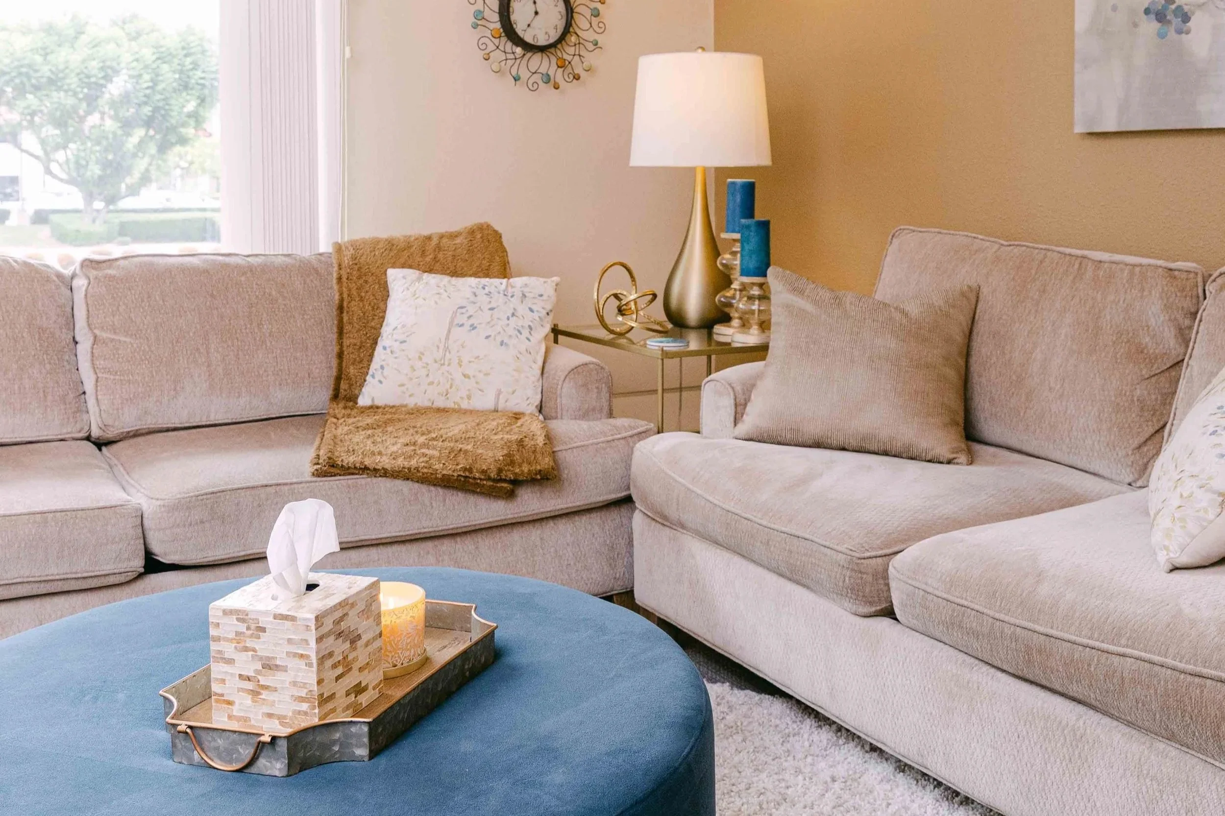 Living room with beige sectional sofas, decorative pillows, a side table with a large lamp, and a wall clock. A blue ottoman holds a tissue box and candles, and a window shows greenery outside.