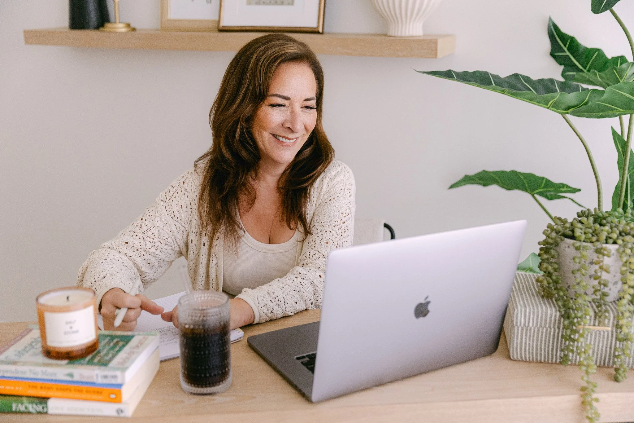 A woman with brown hair smiling while working at a desk with a laptop, notebook, books, a candle, and a glass of dark soda, with plants and framed pictures in the background.