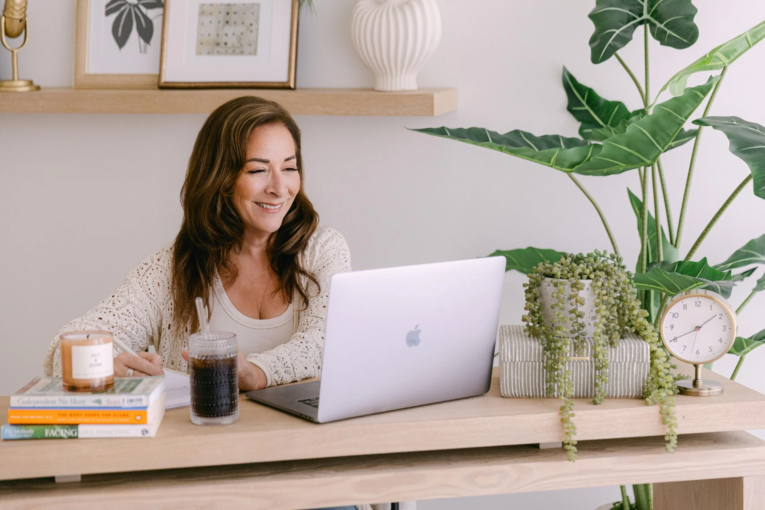 A woman with long brown hair smiling while working on a laptop at a wooden desk, surrounded by books, a candle, a glass of dark beverage, and a large green plant with broad leaves.