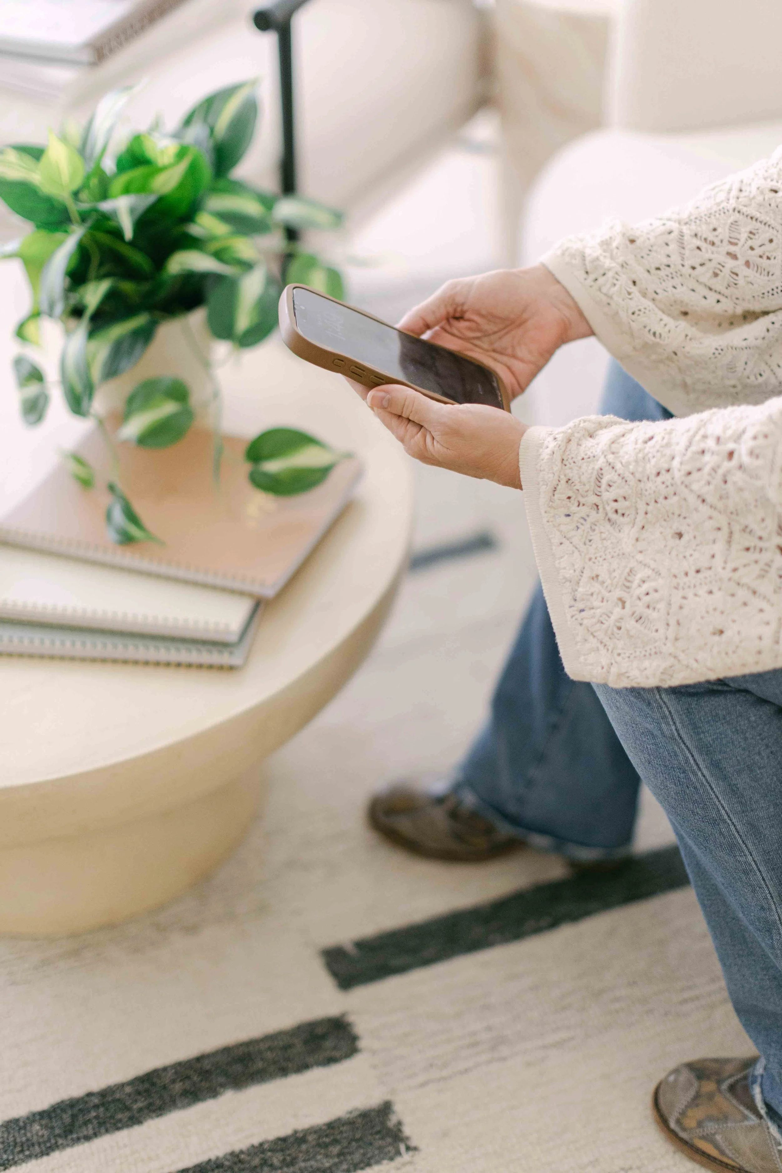 A person in a cream lace top is standing near a round wood table, holding a smartphone. On the table, there is a potted plant with green and white variegated leaves, and some notebooks.