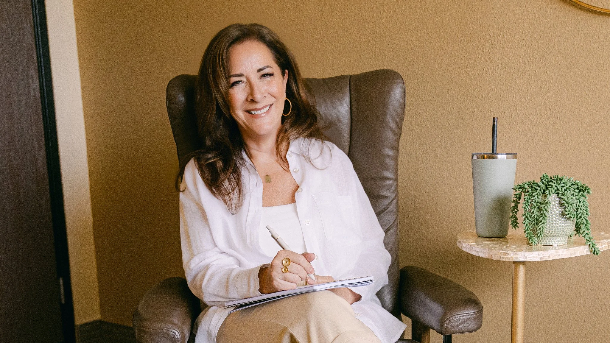 A woman sitting in a brown leather armchair, smiling, holding a pen and notebook in her lap, wearing a white shirt and gold jewelry.