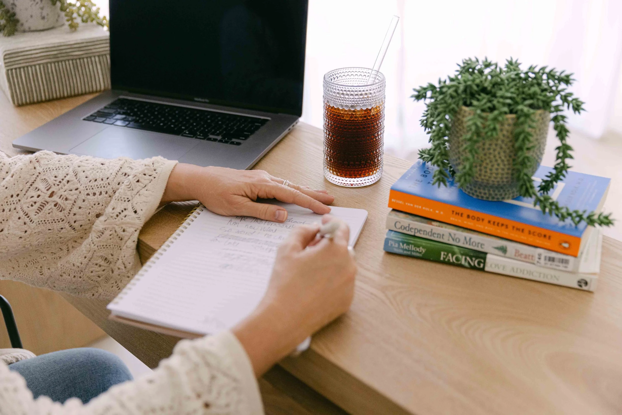 Person taking notes at a wooden desk with a laptop, a glass of iced tea, a potted plant, and a stack of books.