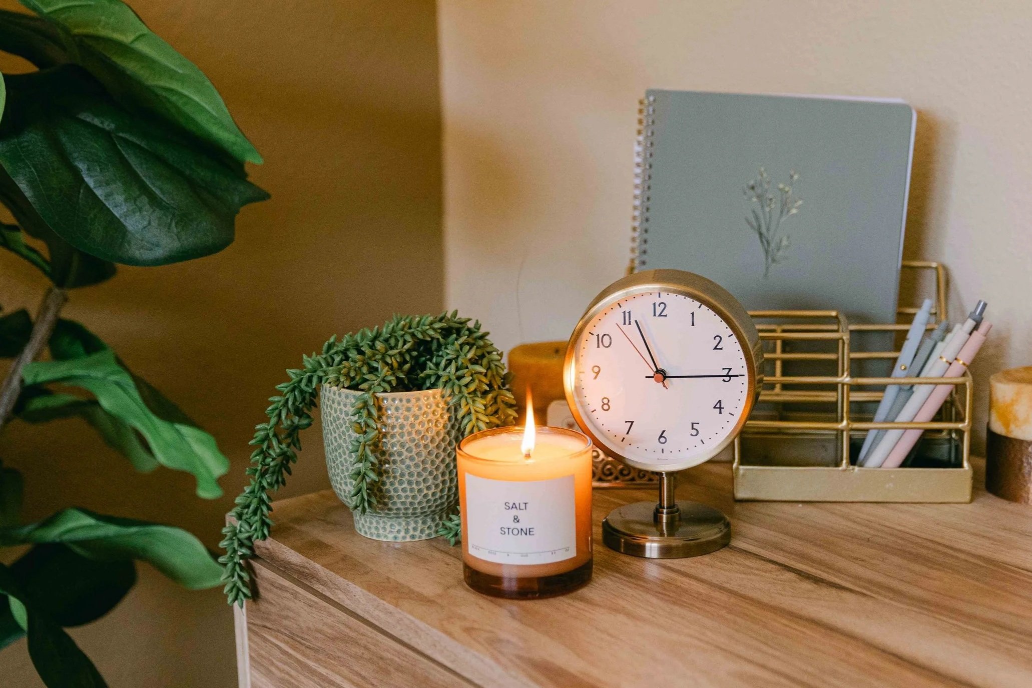 A desk with a potted plant, a lit candle, a white round clock showing approximately 10:56, and a gold-colored wire organizer with pens inside. There is a blue book with a plant illustration and a label that reads "Salt & Stone." The scene is warm and cozy with soft lighting.
