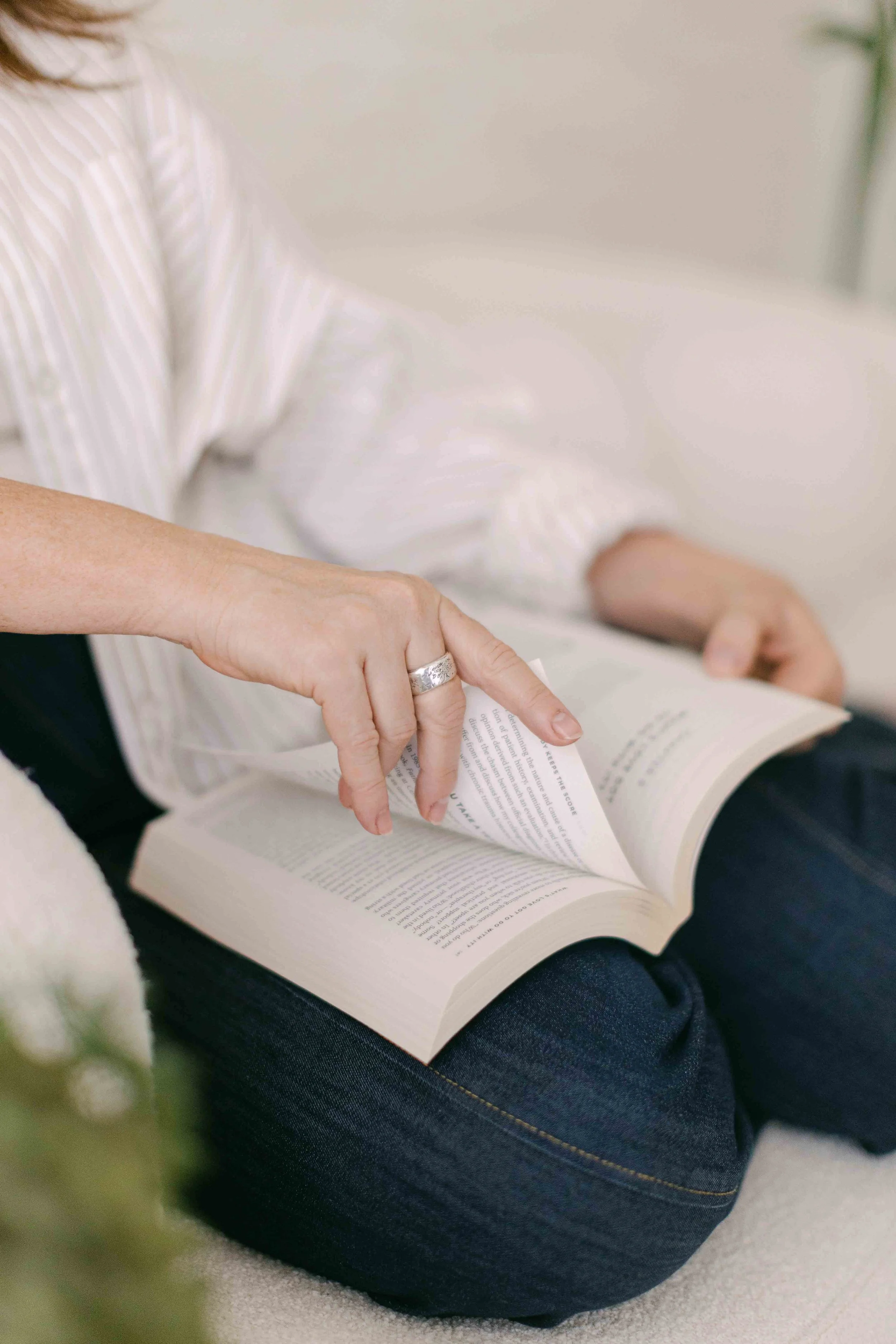 A woman with a silver ring on her finger is reading a book while sitting on a light-colored carpet. She is wearing a white and beige striped shirt and dark jeans.