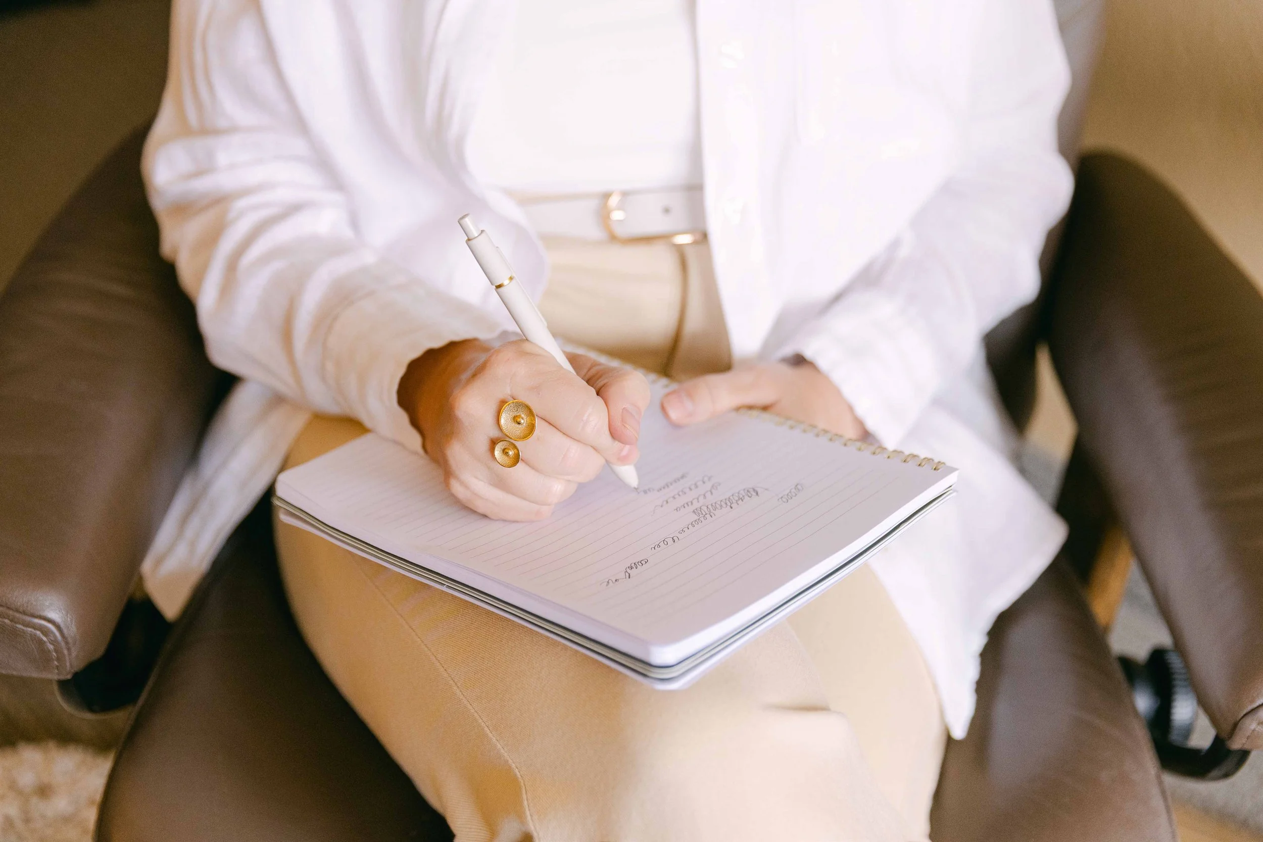 Person wearing a white shirt and tan pants, sitting on a brown chair, holding a white pen, writing in a spiral-bound notebook with handwritten notes.