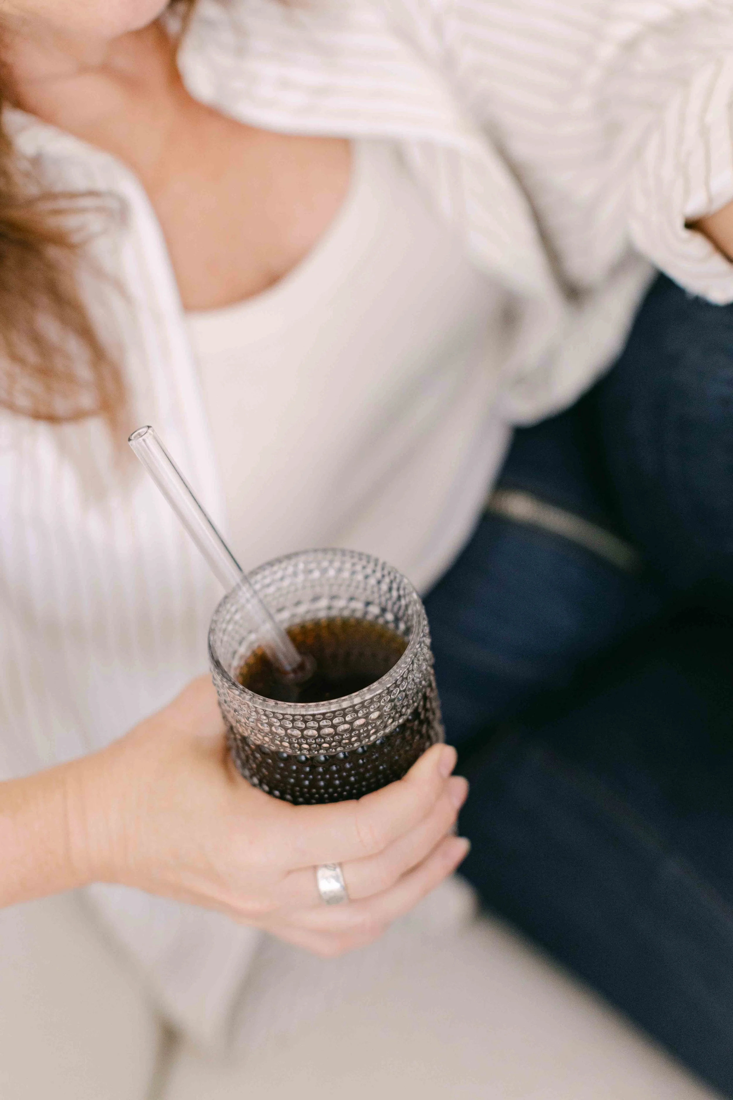 Person holding a glass of soda with a striped straw, wearing a white top and dark pants.