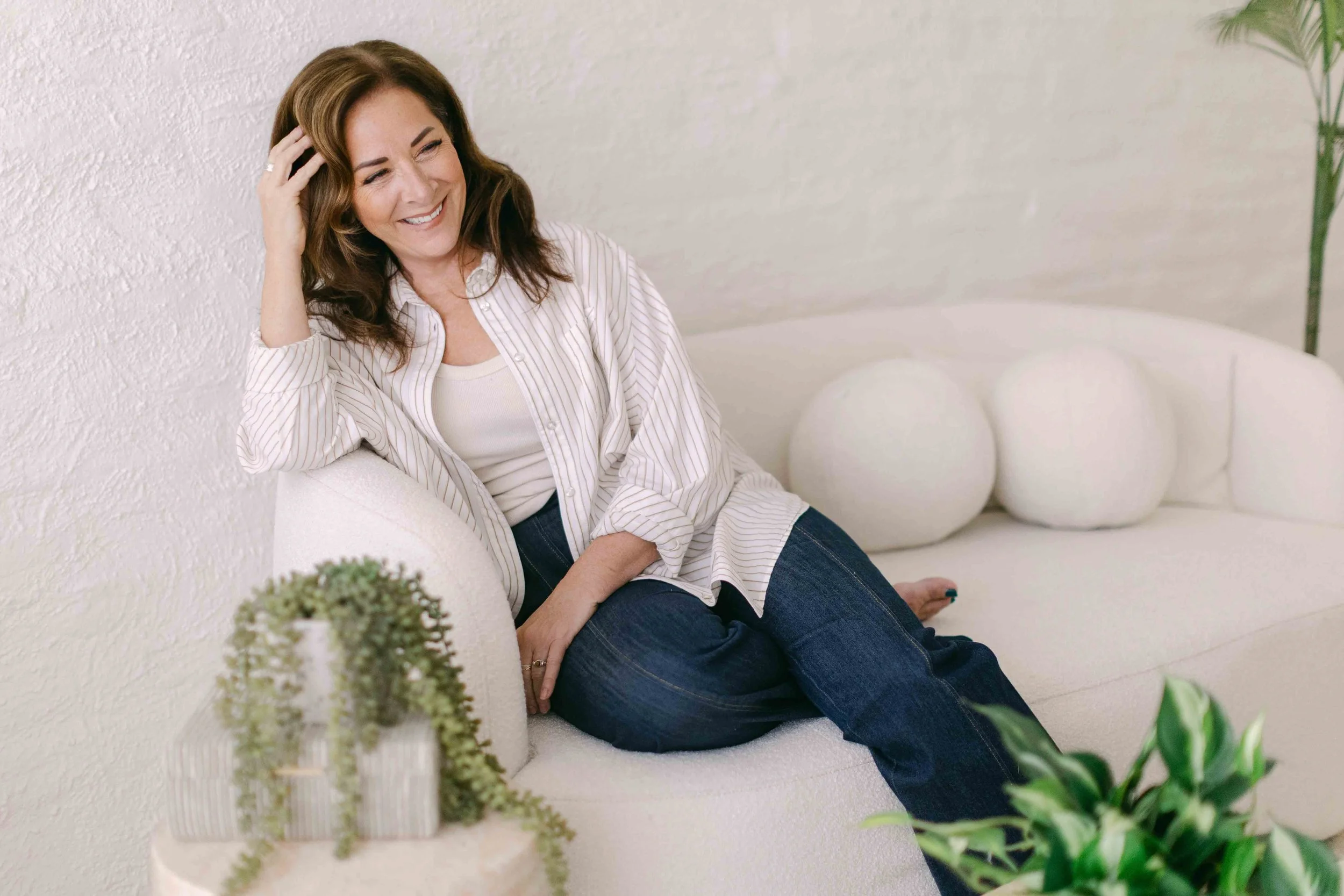 Woman with brown hair smiling, sitting on a white sofa in a bright room decorated with plants.