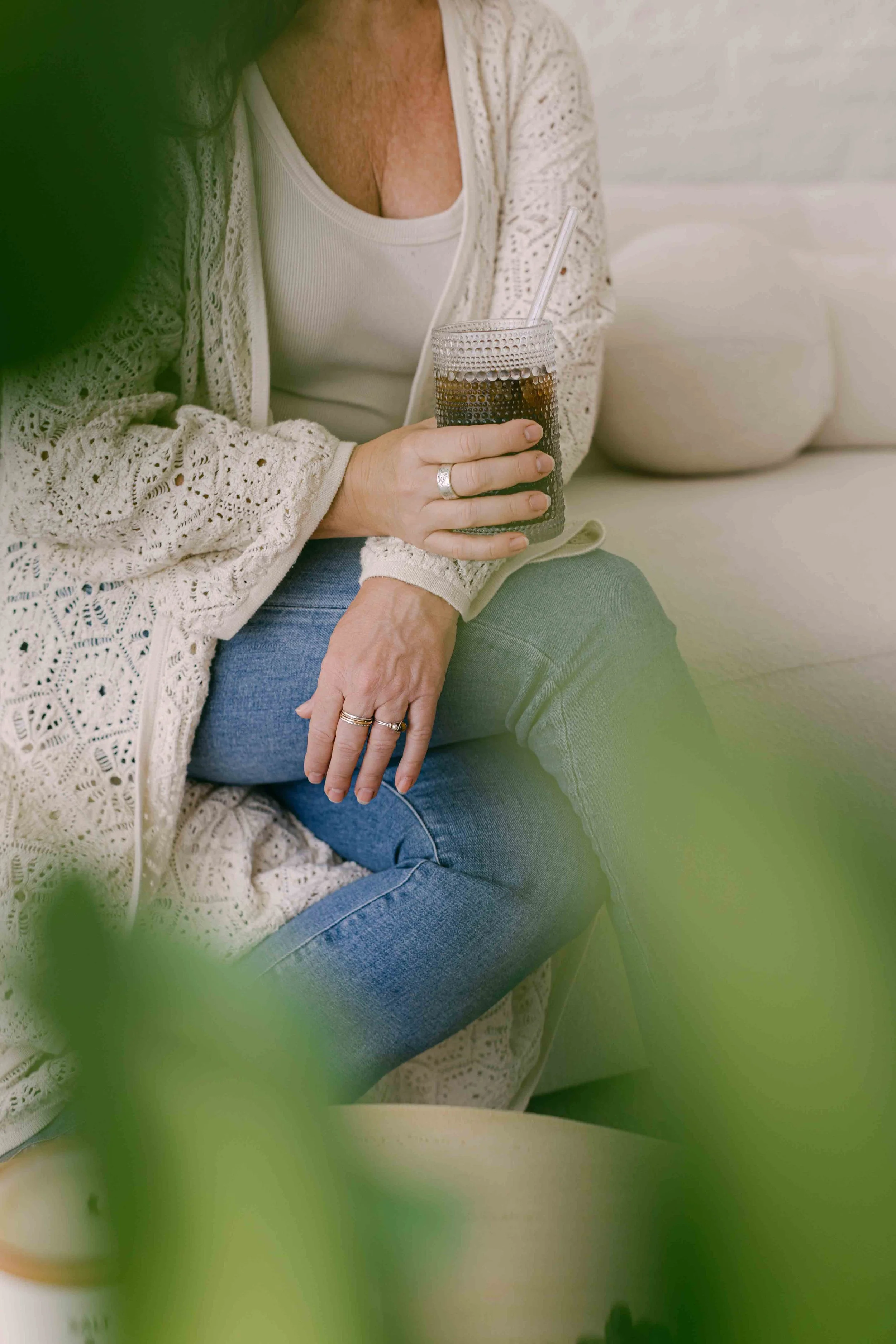 A woman holding a glass of cola with a straw. She is wearing a white tank top and a cream-colored, lace-patterned jacket, sitting on a light-colored couch. There are some green leaves in the foreground, partially obscuring the view.