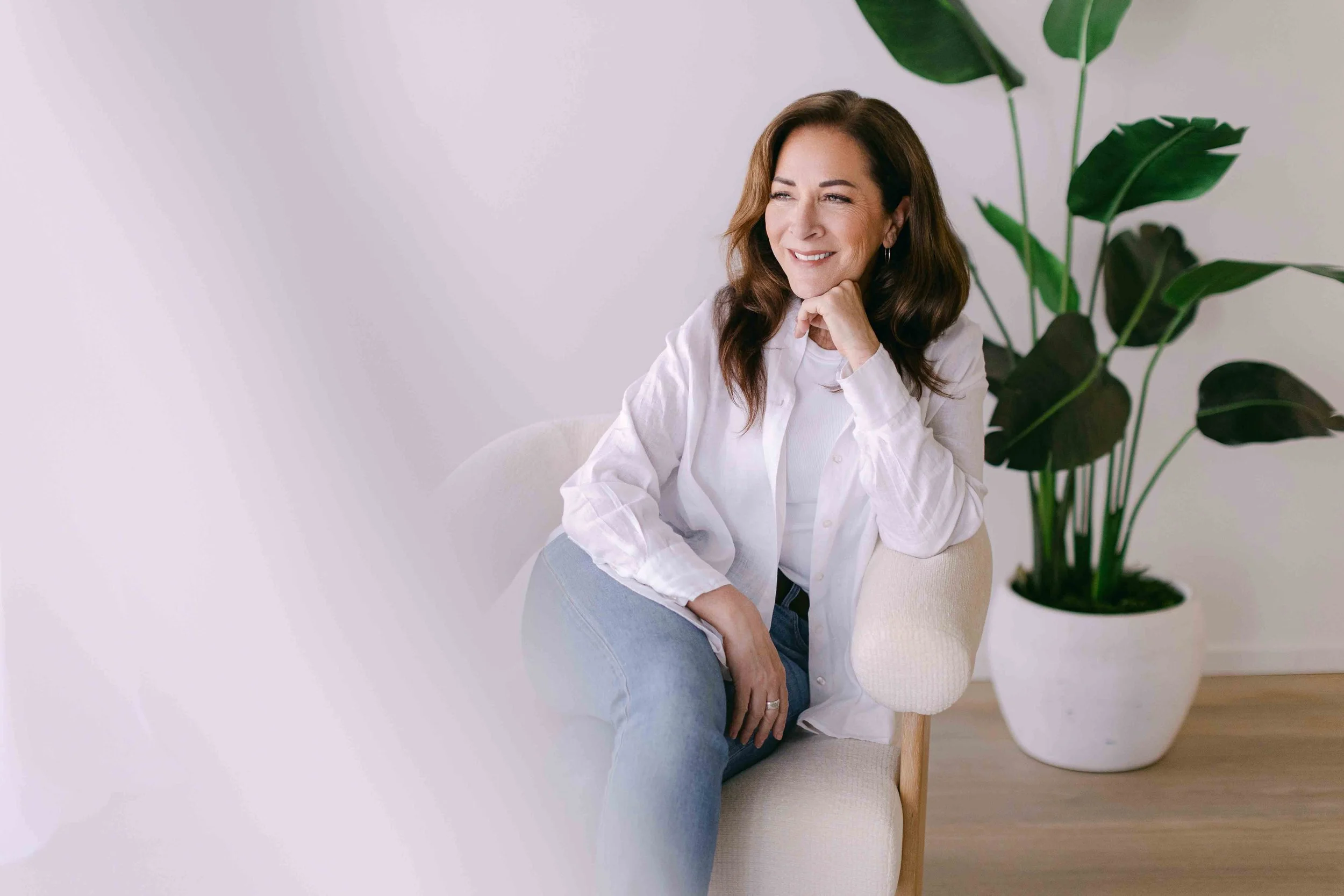 A woman with shoulder-length brown hair smiling, sitting on a light-colored chair with her chin resting on her hand, next to a large potted plant with dark green leaves, in a room with a white wall background.