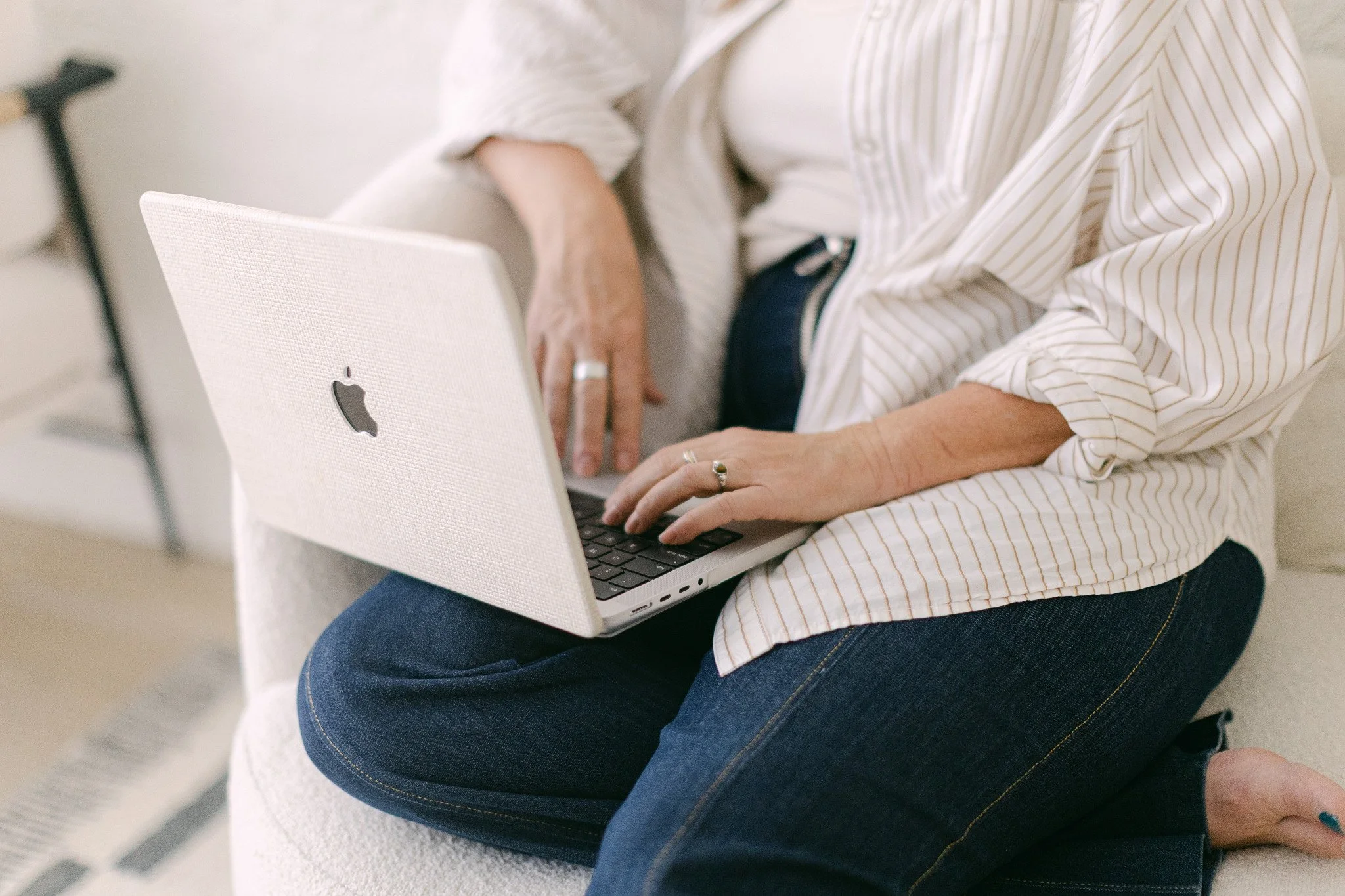 Person sitting on a white chair using a MacBook with a white textured cover, wearing a white striped shirt and blue jeans.