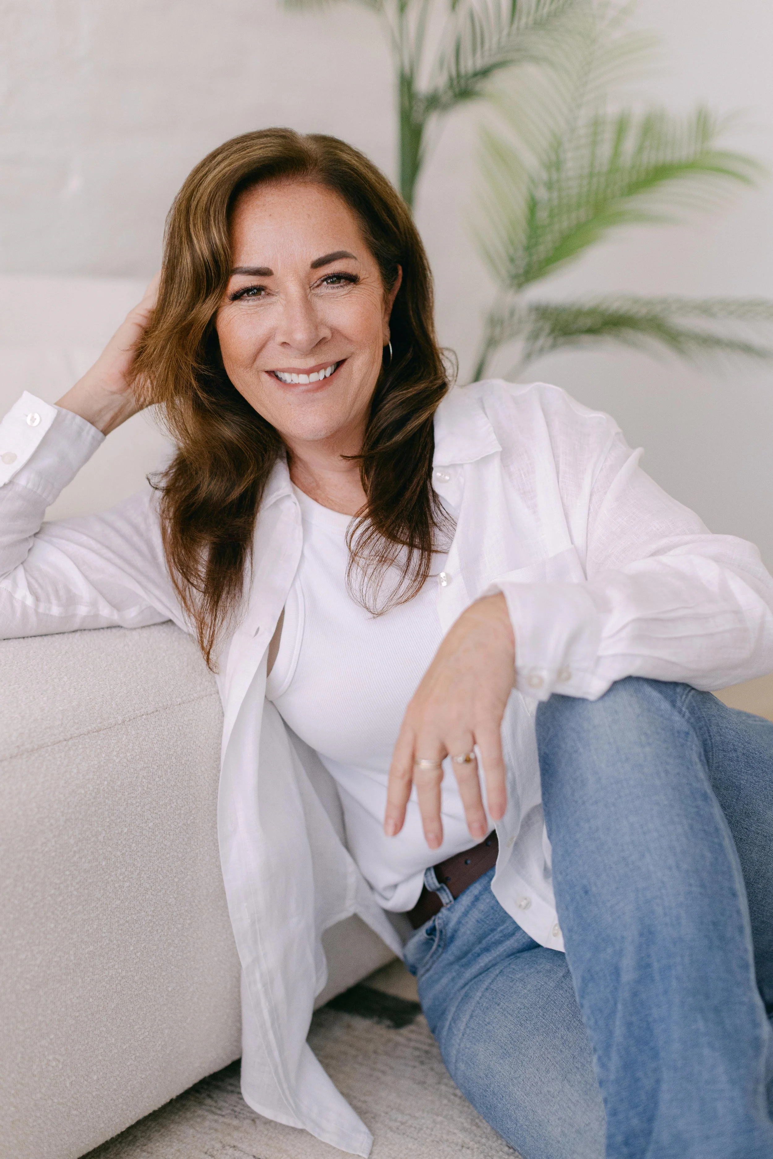 A woman with brown hair and a white shirt smiling while sitting on a beige couch, with a man in blue jeans lying on her lap, in a room with a potted plant in the background.
