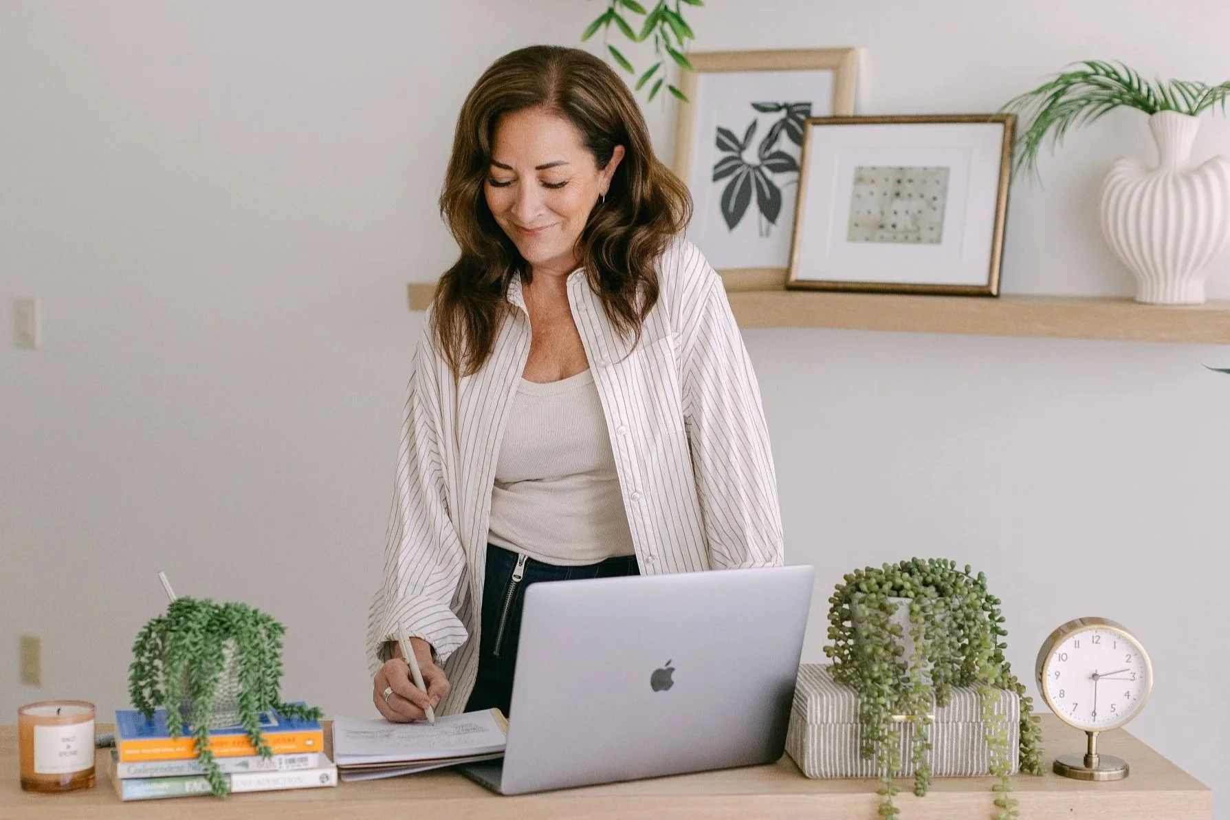 A woman with long brown hair, wearing a white striped shirt, working at a desk with a laptop, writing on a notepad. The desk has green plants, a clock, and books.