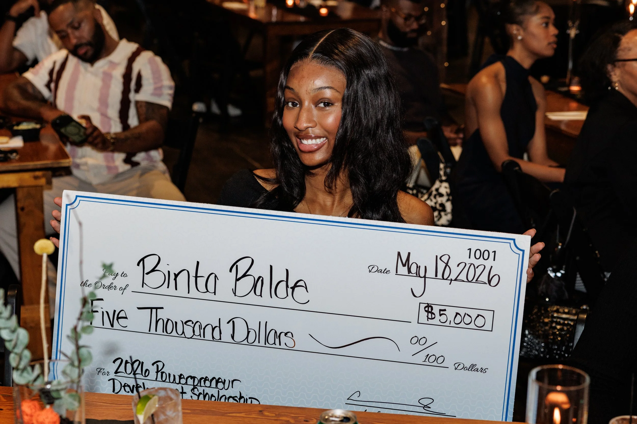 A young woman with long dark hair holding a large check for five thousand dollars at an indoor event, with other people seated at tables in the background.