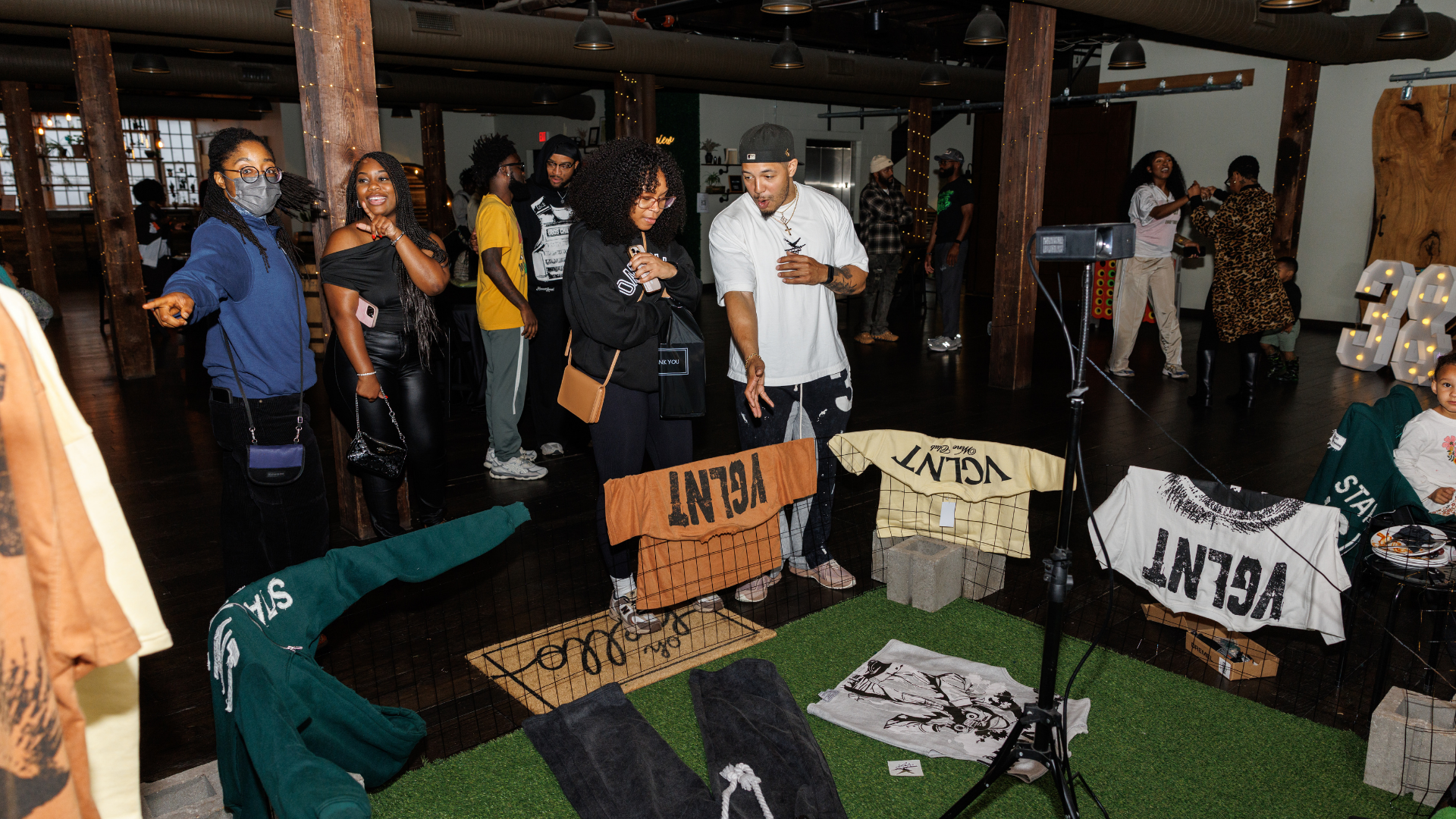A group of people at an indoor event, standing behind a display with various clothing and merchandise. Some are looking at the display while others are socializing or dancing in the background. The setting has warm lighting and a cozy atmosphere.
