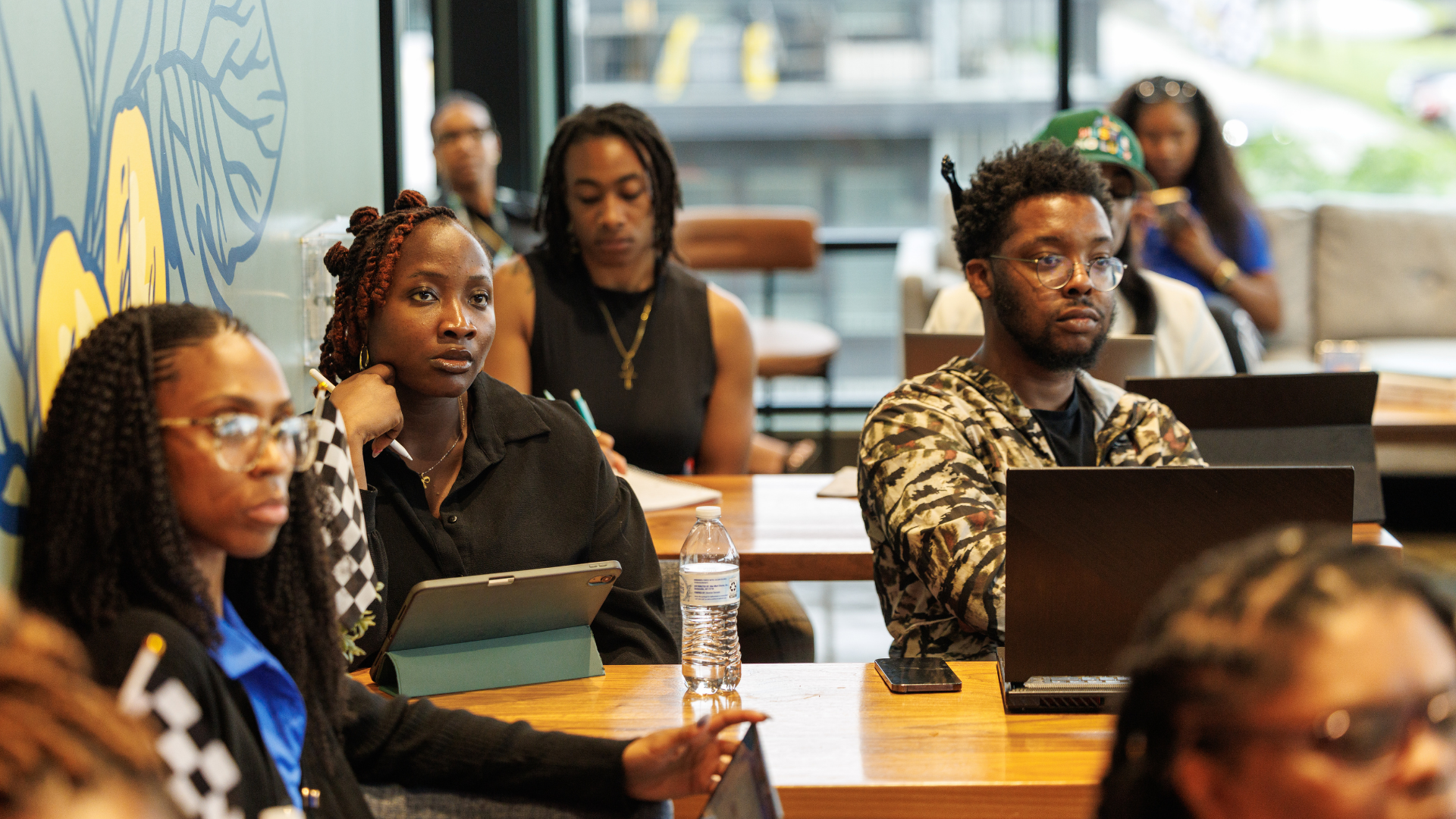 People attending a meeting or class in a modern room with large windows, some with laptops and tablets.
