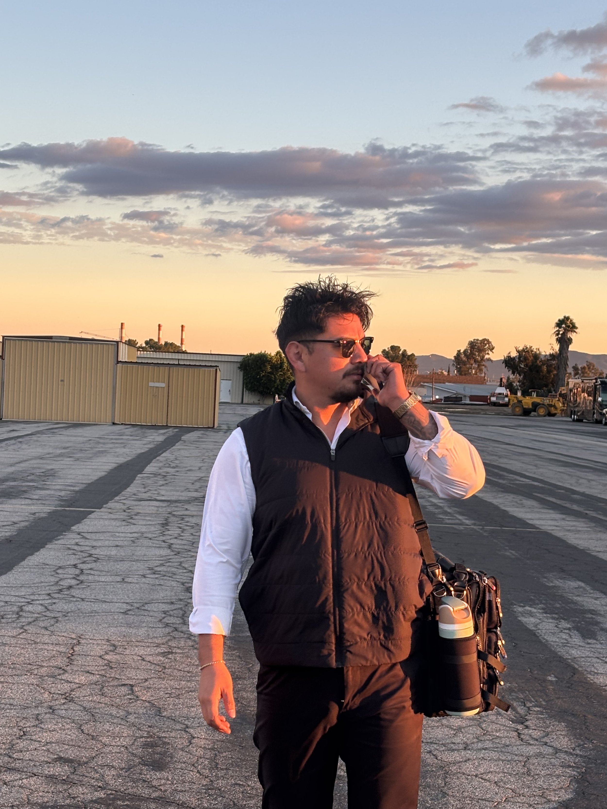 A man with dark hair, sunglasses, and a mustache stands outdoors at sunset, holding a water bottle and adjusting his sunglasses. He is dressed in a white shirt and a black vest, with a camera strap around his neck, in a parking lot with industrial buildings and trees in the background.