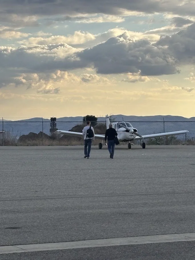 Two people walking towards a parked small white airplane on an airport tarmac during cloudy sunset.