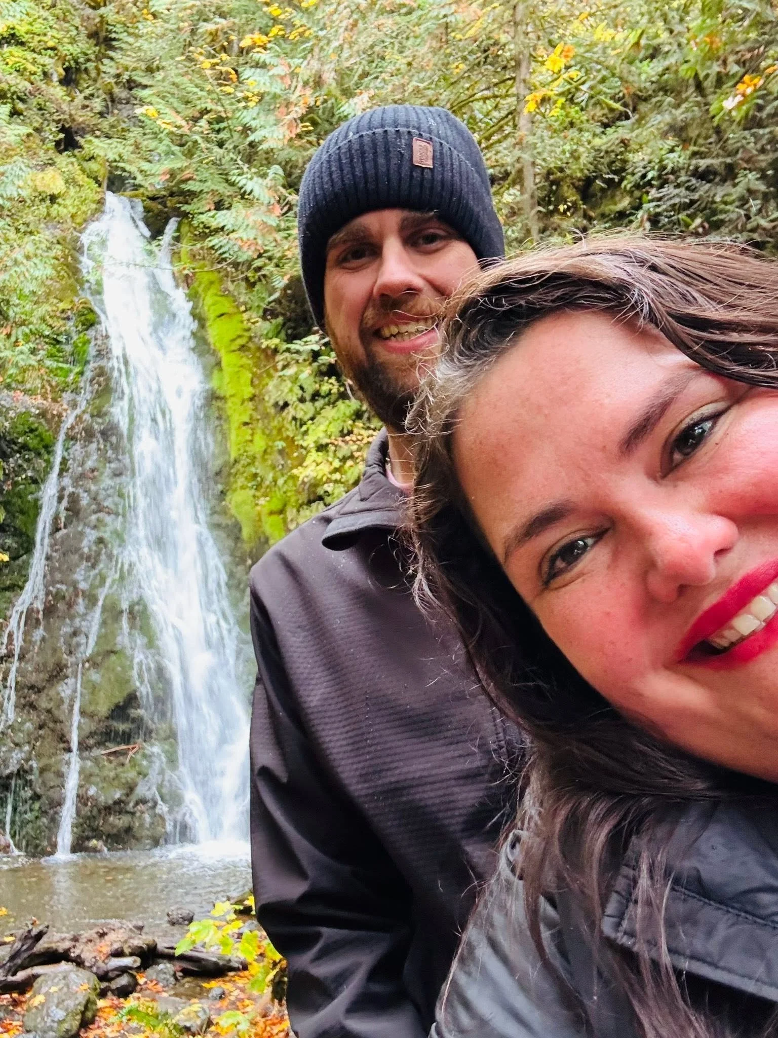 Two smiling people taking a selfie in front of a waterfall surrounded by autumn foliage in a forest.