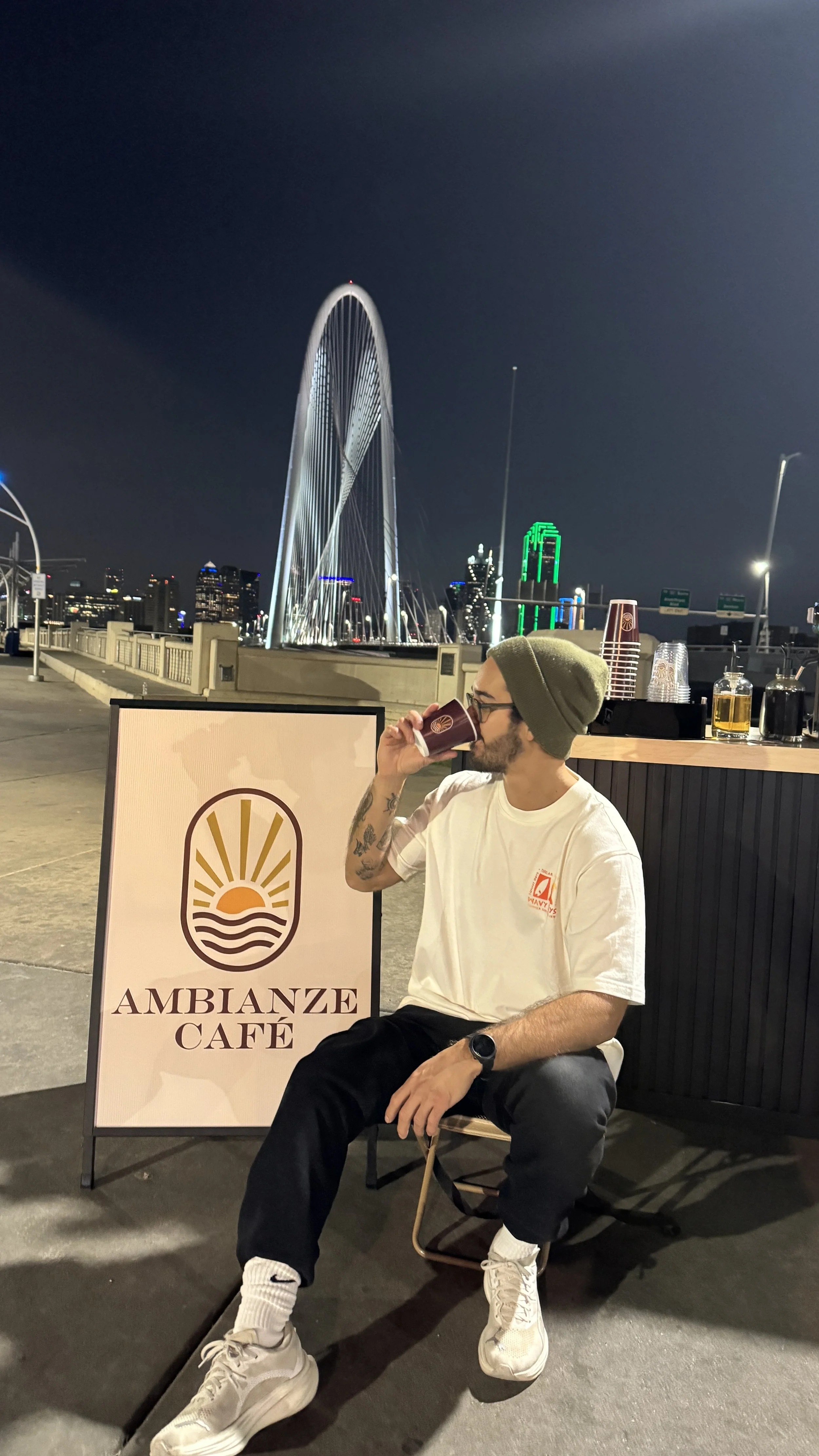 A man with tattoos wearing a gray beanie, white T-shirt, and black pants sitting outside at night, sipping from a cup at Ambanze Café with a city skyline and illuminated buildings in the background.