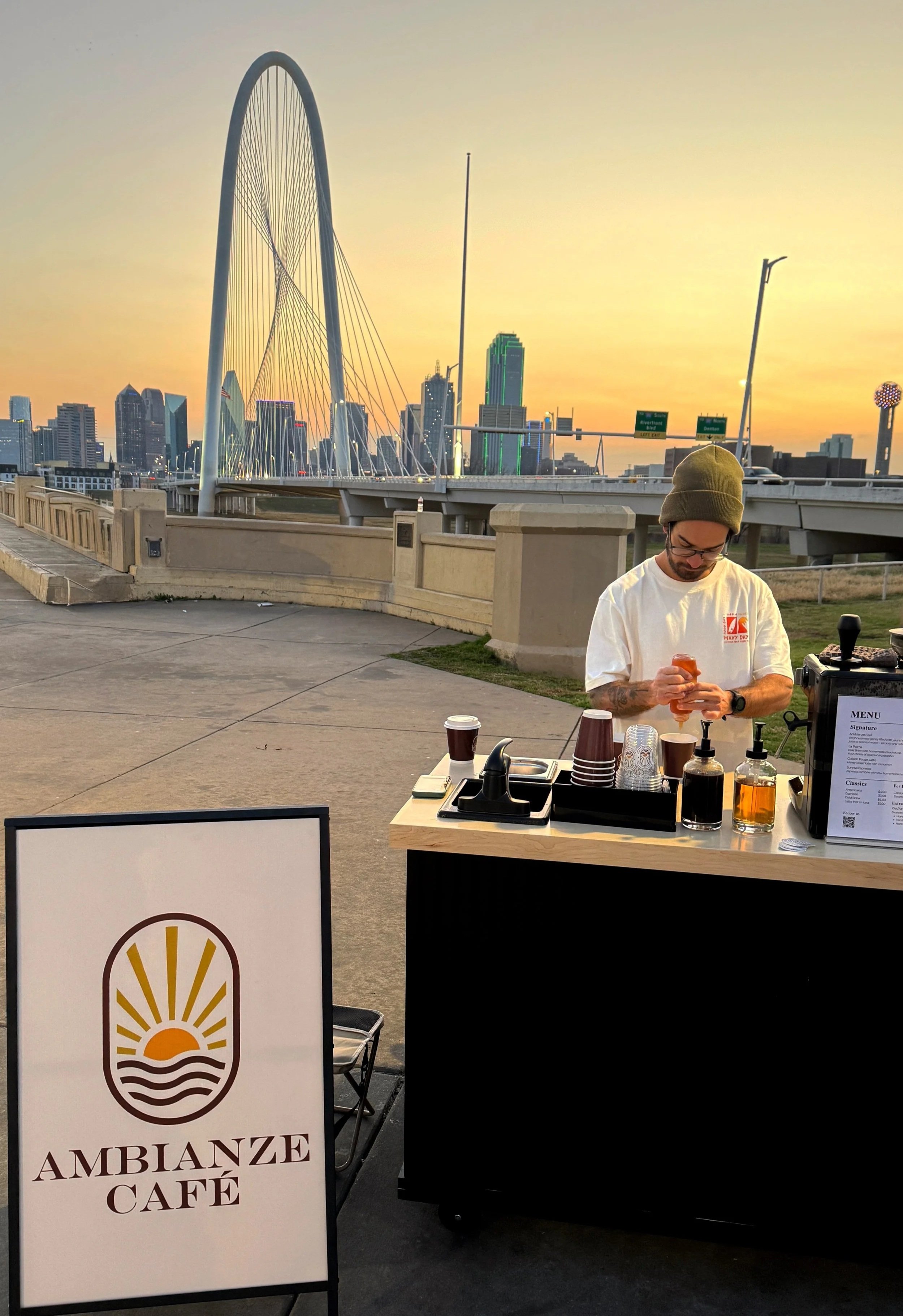 A man wearing a green beanie and glasses working at a coffee stand with a sign that reads 'Ambianze Café' in front of a city skyline at sunset, including a large arch bridge.