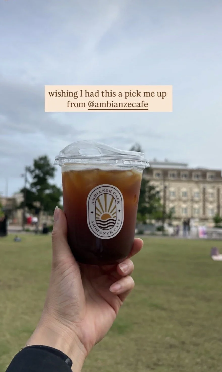 A hand holds a clear plastic cup filled with iced coffee from Ambianze Cafe, with a cafe logo on the cup, in an outdoor park setting with a large building and trees in the background.
