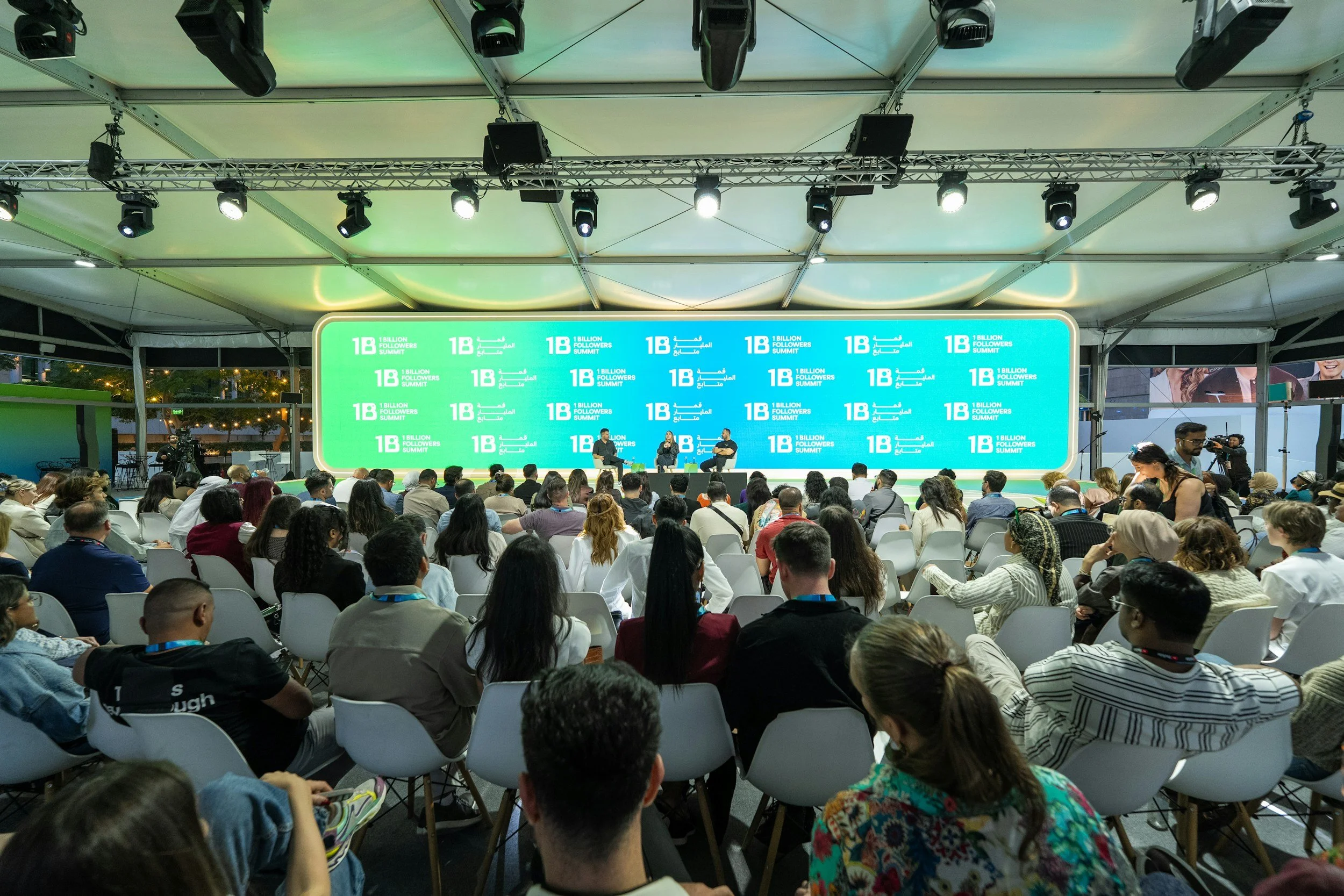 A crowded conference or seminar with people seated and a large digital screen on stage displaying event branding for the '1 Billion Followers Summit'.
