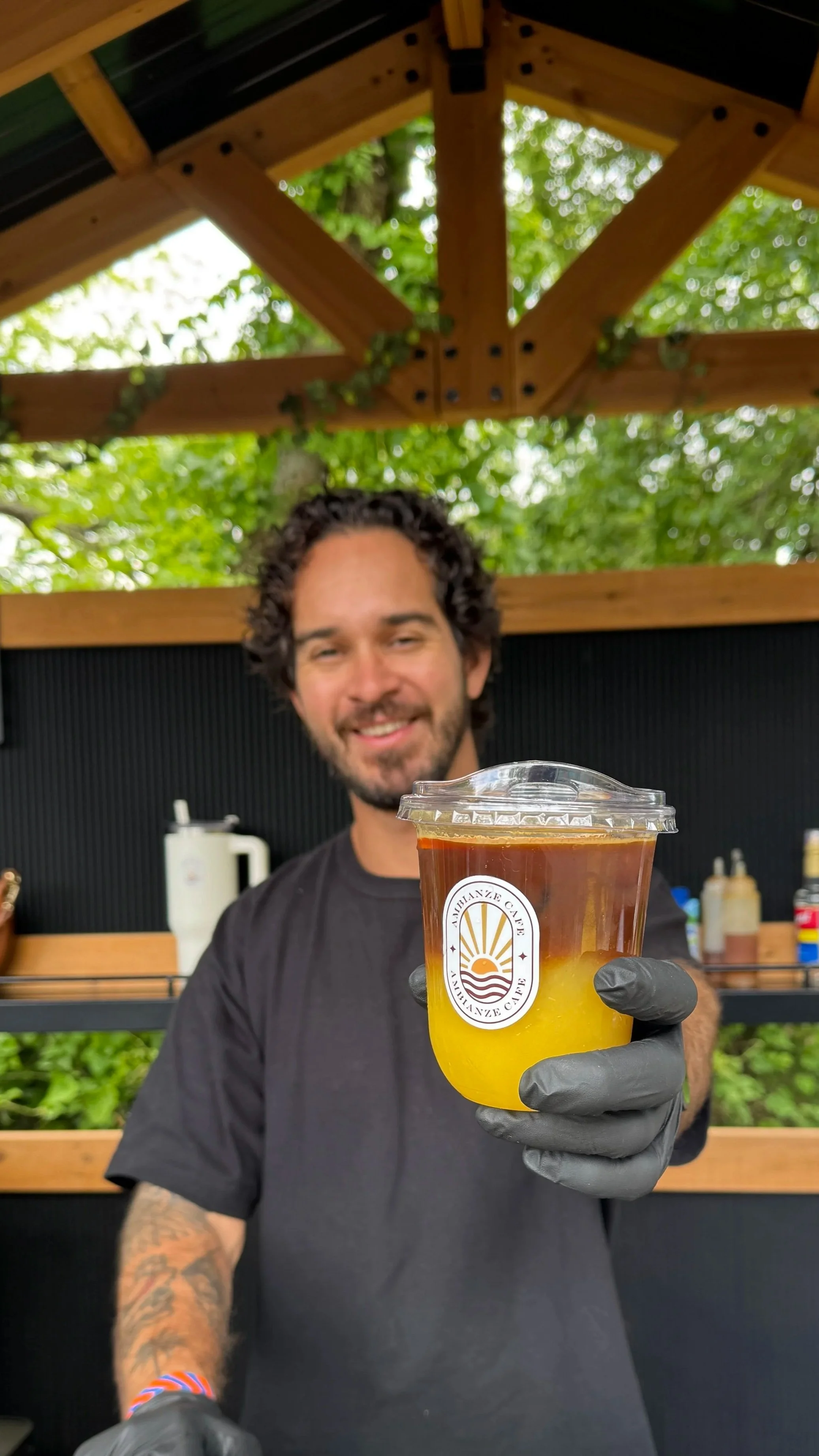 A smiling man with curly dark hair, a beard, and tattoos on his arm, wearing a black t-shirt and grey gloves, holding a plastic cup of yellow and dark brown layered beverage outside under a wooden structure with green leaves in the background.