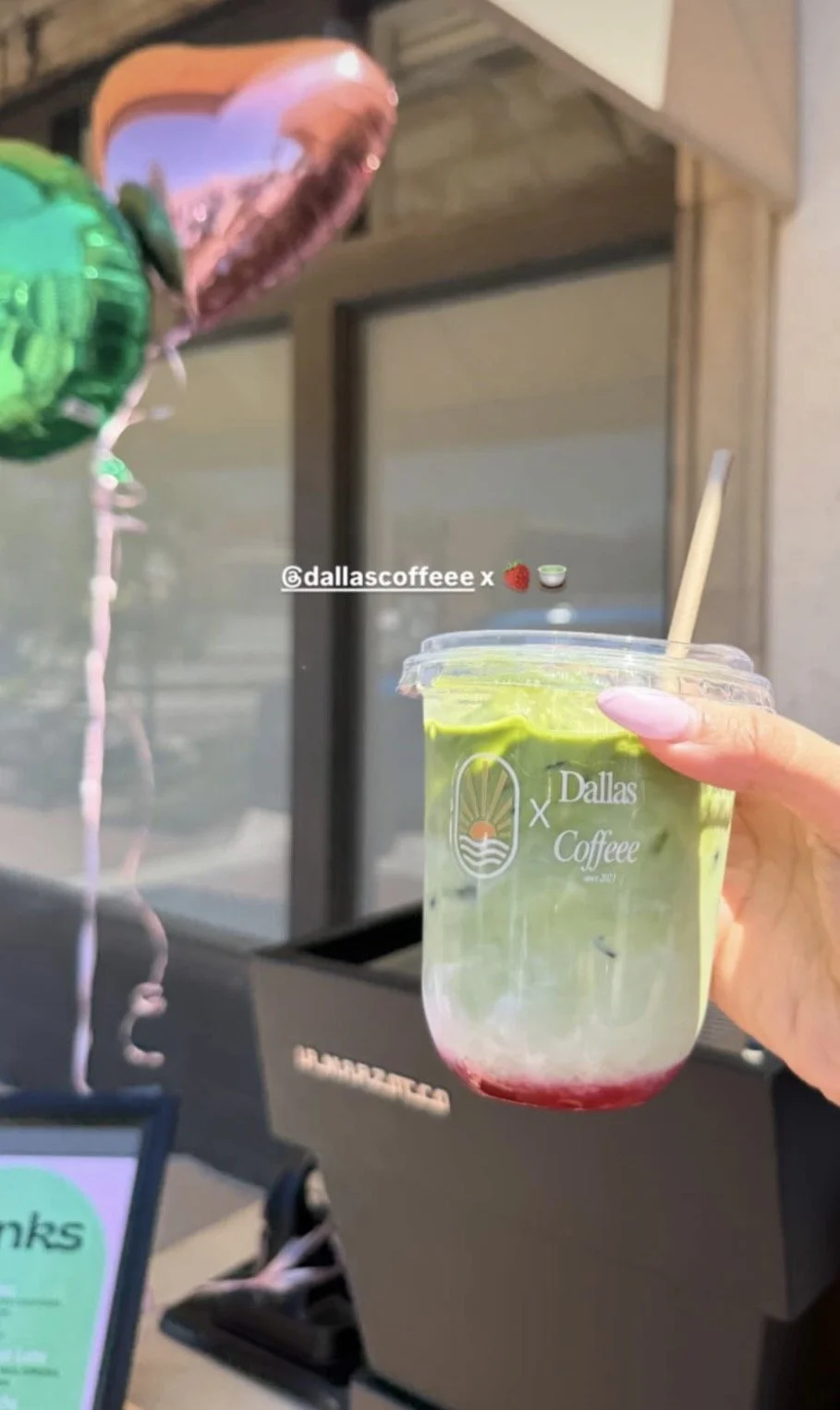 A hand holding a clear plastic cup with a green drink, red syrup at the bottom, and a wooden stir stick. The cup has the 'Dallas Coffee' logo. In the background, there are pink, green, and rose gold balloons, and a drive-thru window.