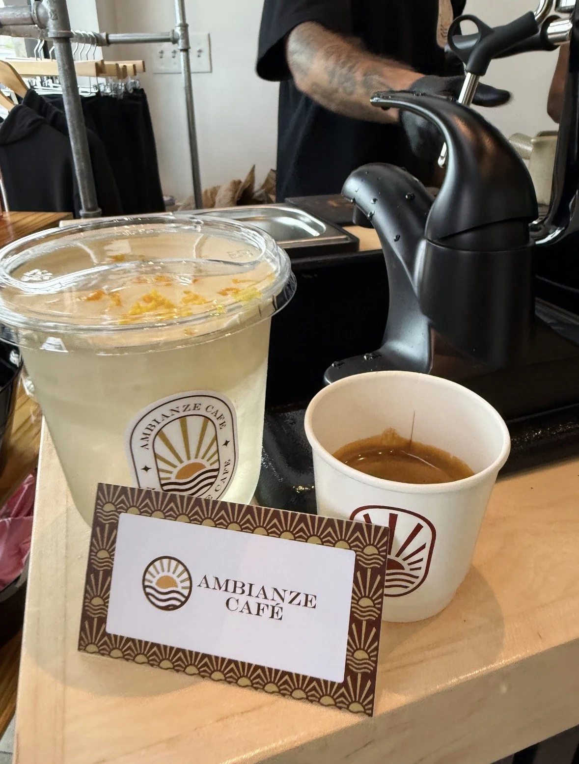 A coffee shop scene at Ambianze Café showing two cups of coffee, one larger and one smaller, on a wooden table. In the background, there is a coffee grinder, a person wearing a black shirt operating it, and clothing racks with black garments nearby.