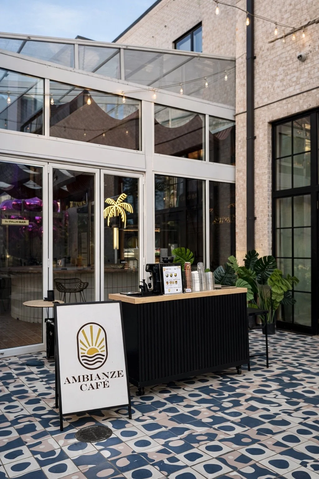 Outdoor café with a sign reading 'Ambianze Café', a coffee station with cups and beverage dispensers, black and white patterned tile floor, glass walls, green plants, and string lights overhead.