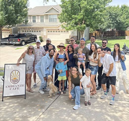 Group of people gathering outdoors in front of a house, some holding drinks, with a sign for Ambianze Cafe, and a dog on a leash.