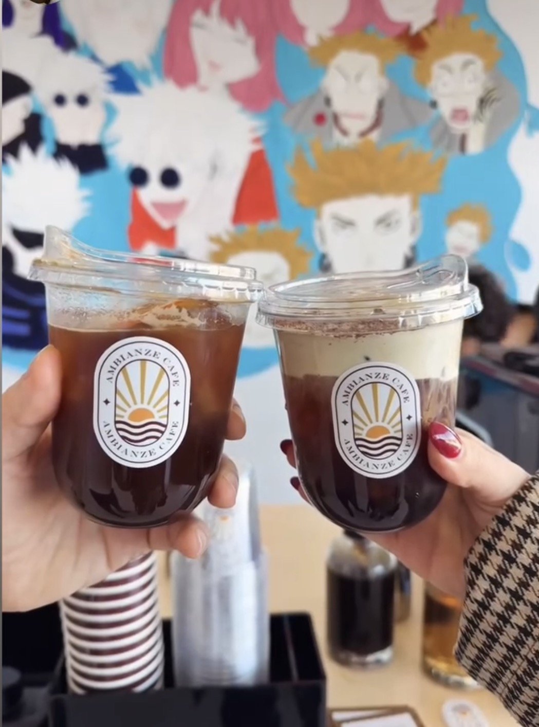 Two hands holding iced coffee drinks with a logo, in front of a colorful mural with various cartoon characters.