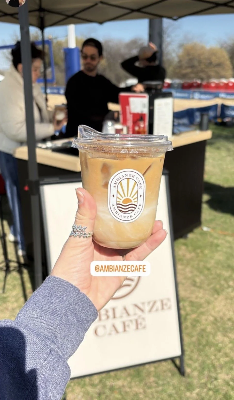 A person holding iced coffee with ice cubes and a straw in front of a cafe stand, with two people in the background under a canopy at an outdoor event.