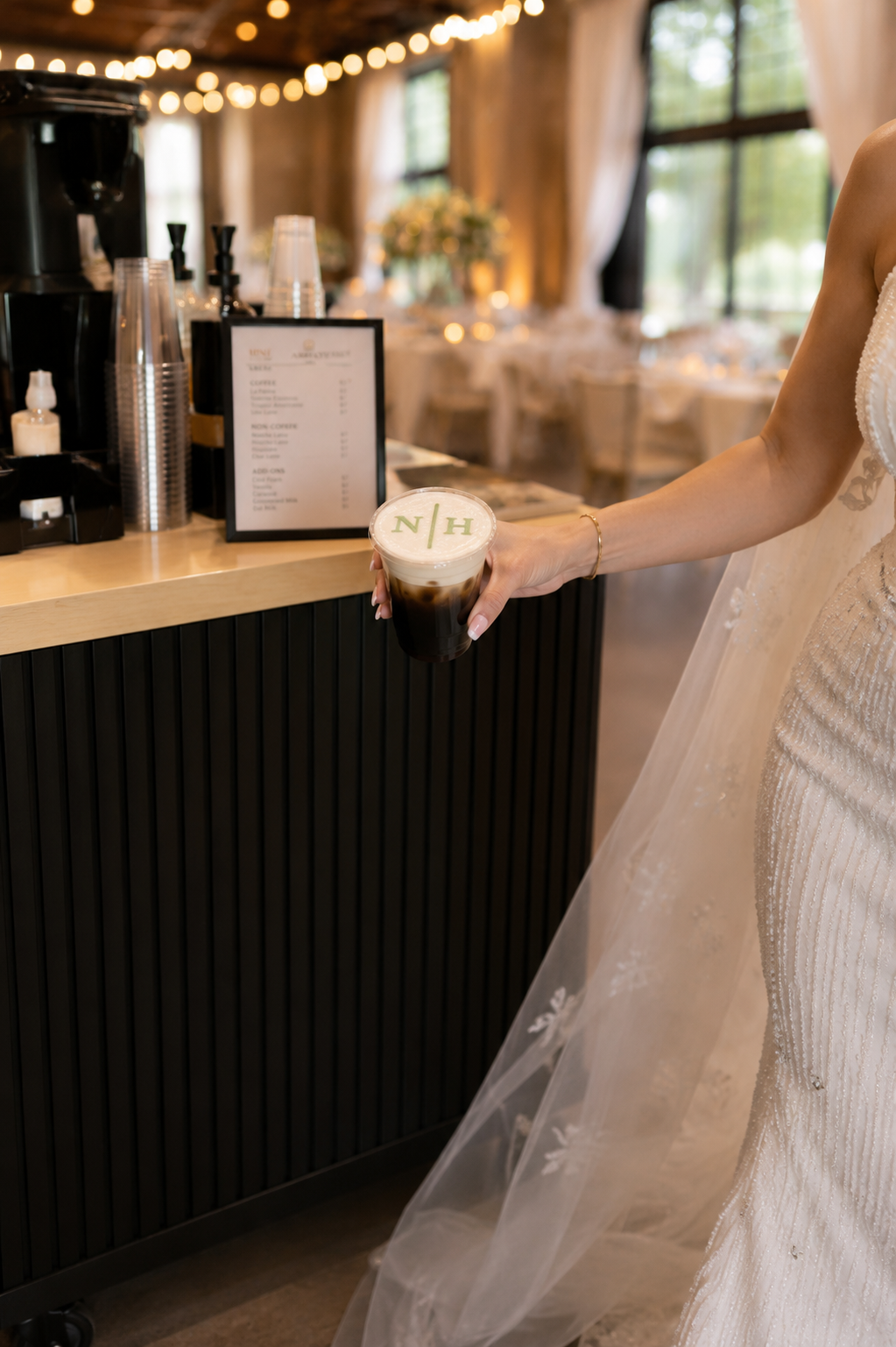 A person in a wedding dress holding a cup of iced coffee with green Starbucks logo on the foam in a cozy cafe with string lights and large windows.