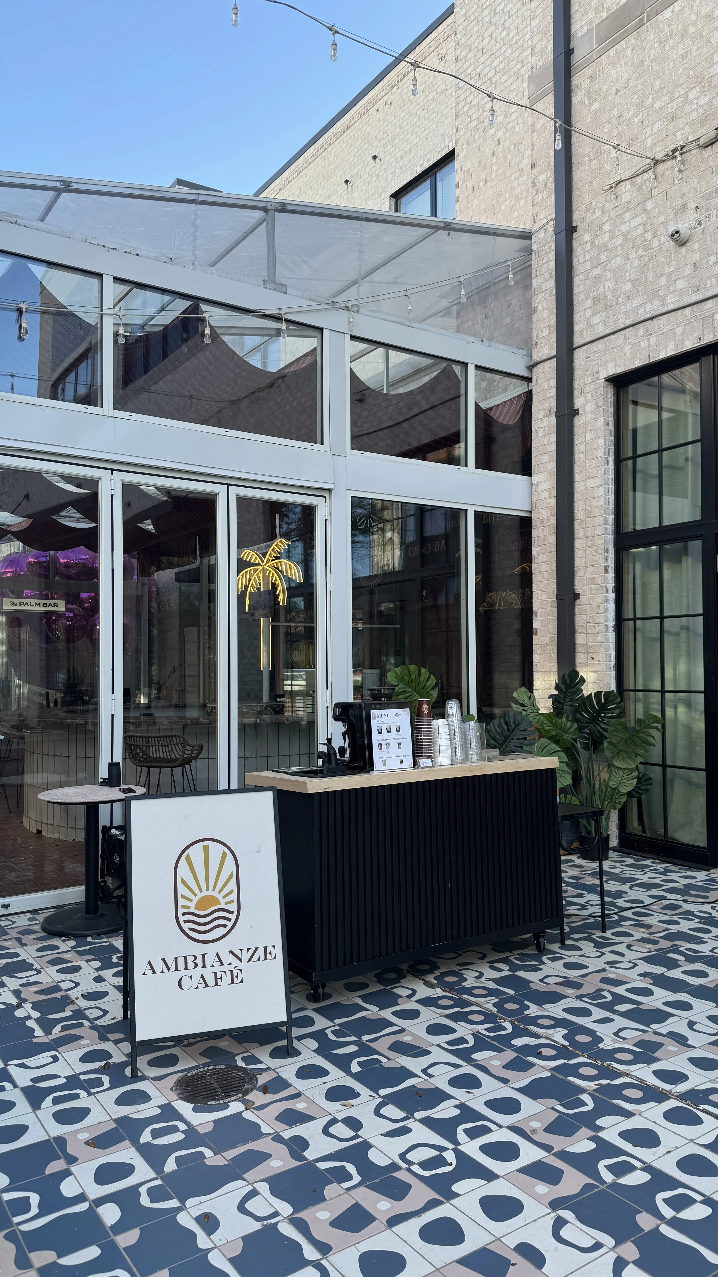 Outdoor view of Ambianze Café with a signboard, serving area with a coffee machine, cups, and decorative green plants, on a patterned tiled floor, with a glass-enclosed seating area and string lights overhead, in daytime.