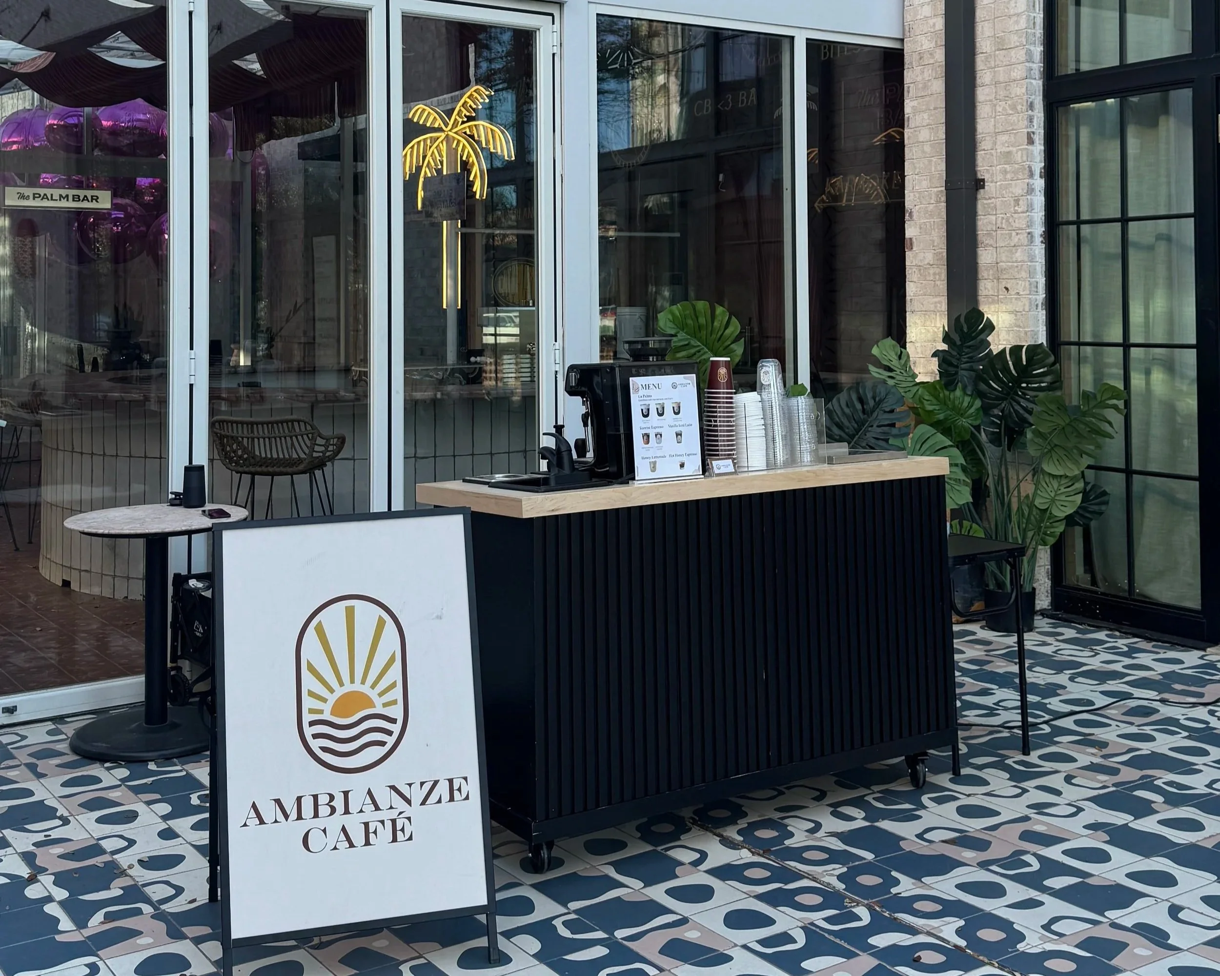 Outside view of a café named Ambanze Café with a neon palm tree sign on the glass door. There are stacked disposable coffee cups, a menu stand, and seating with wicker chairs inside. The floor has patterned blue, white, and beige tiles, and large green plants are visible near the entrance.