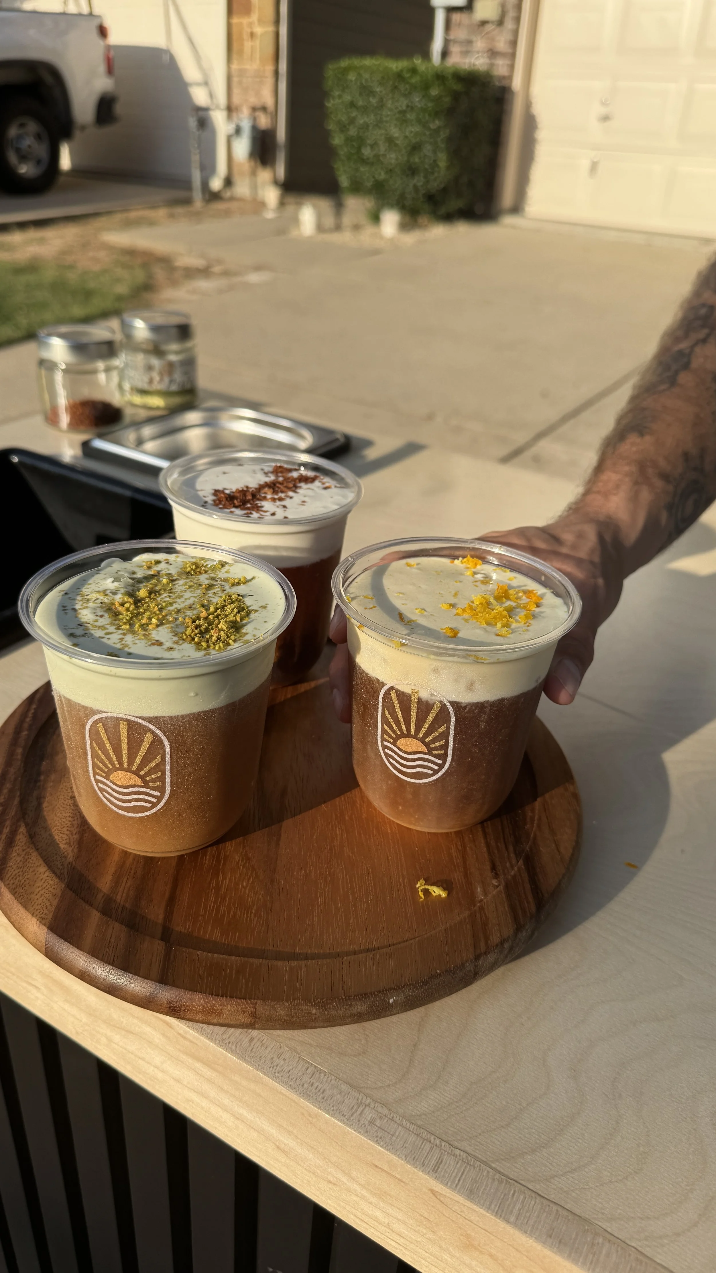 Three beverages with foam and toppings on a round wooden tray, held by a tattooed hand, indoors with a driveway and garage door in the background.