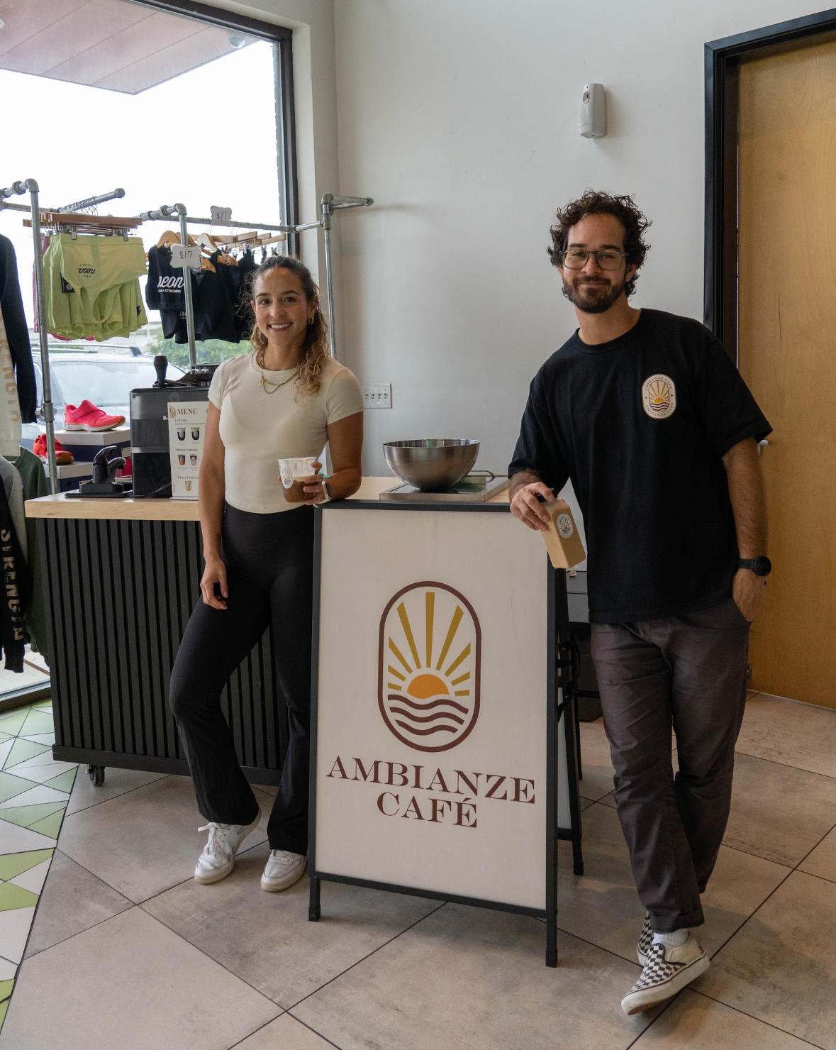 A woman and a man standing behind a café counter at Ambianze Café, smiling. The woman is holding a drink, and there are clothing items on racks in the background.