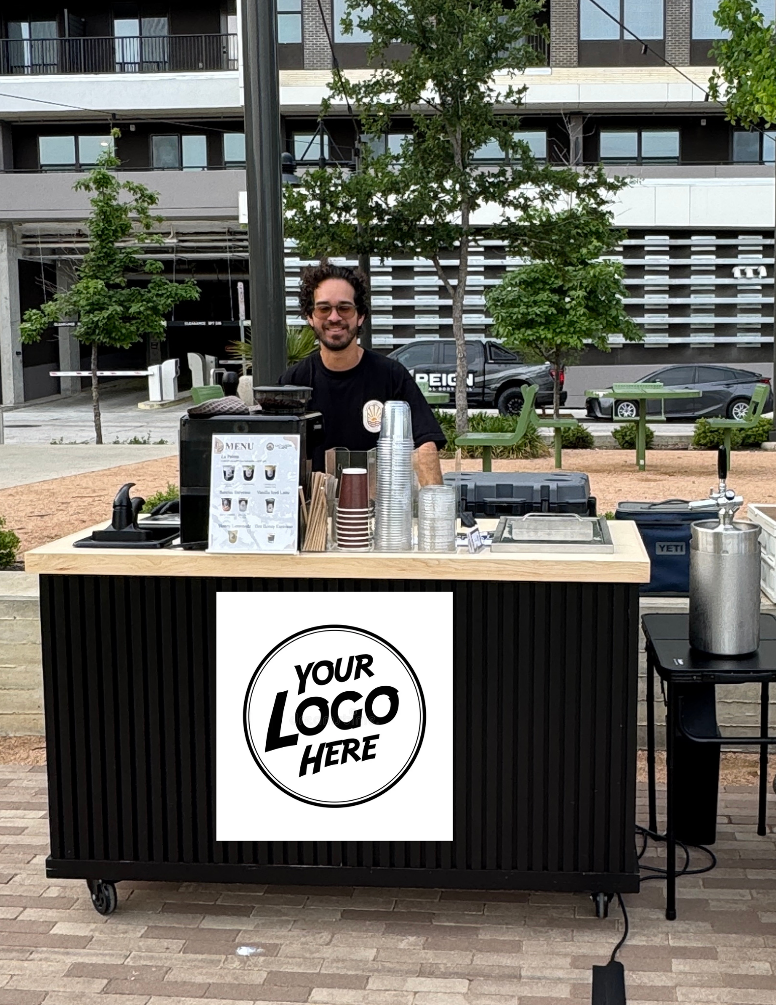 A man with glasses smiling behind a mobile coffee stand with containers, cups, and a menu, in an urban outdoor setting with trees, cars, and modern buildings in the background.