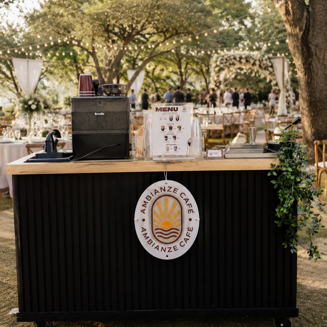 A coffee and beverage stand at an outdoor event with a sign that reads 'AMBIANZE CAFÉ'. In the background, there are tables, chairs, and people gathered under string lights in a park surrounded by trees.
