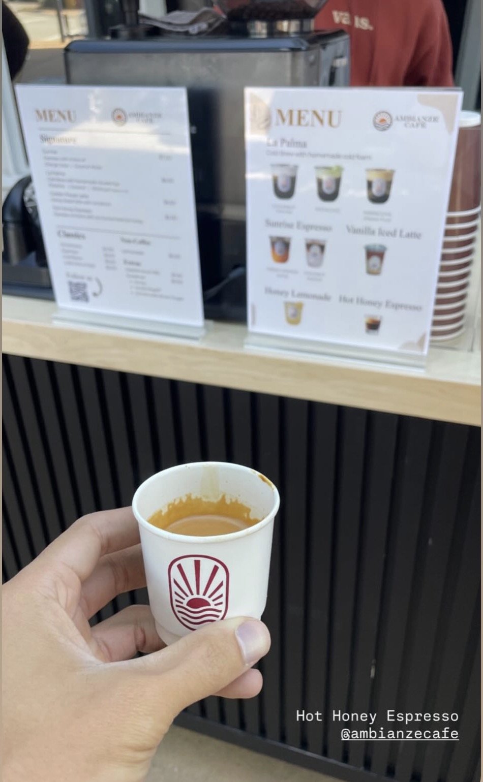 A person holding a small paper cup with the Ambianze Cafe logo, containing hot honey espresso. In the background, there is a counter with menu signs and a commercial coffee machine.